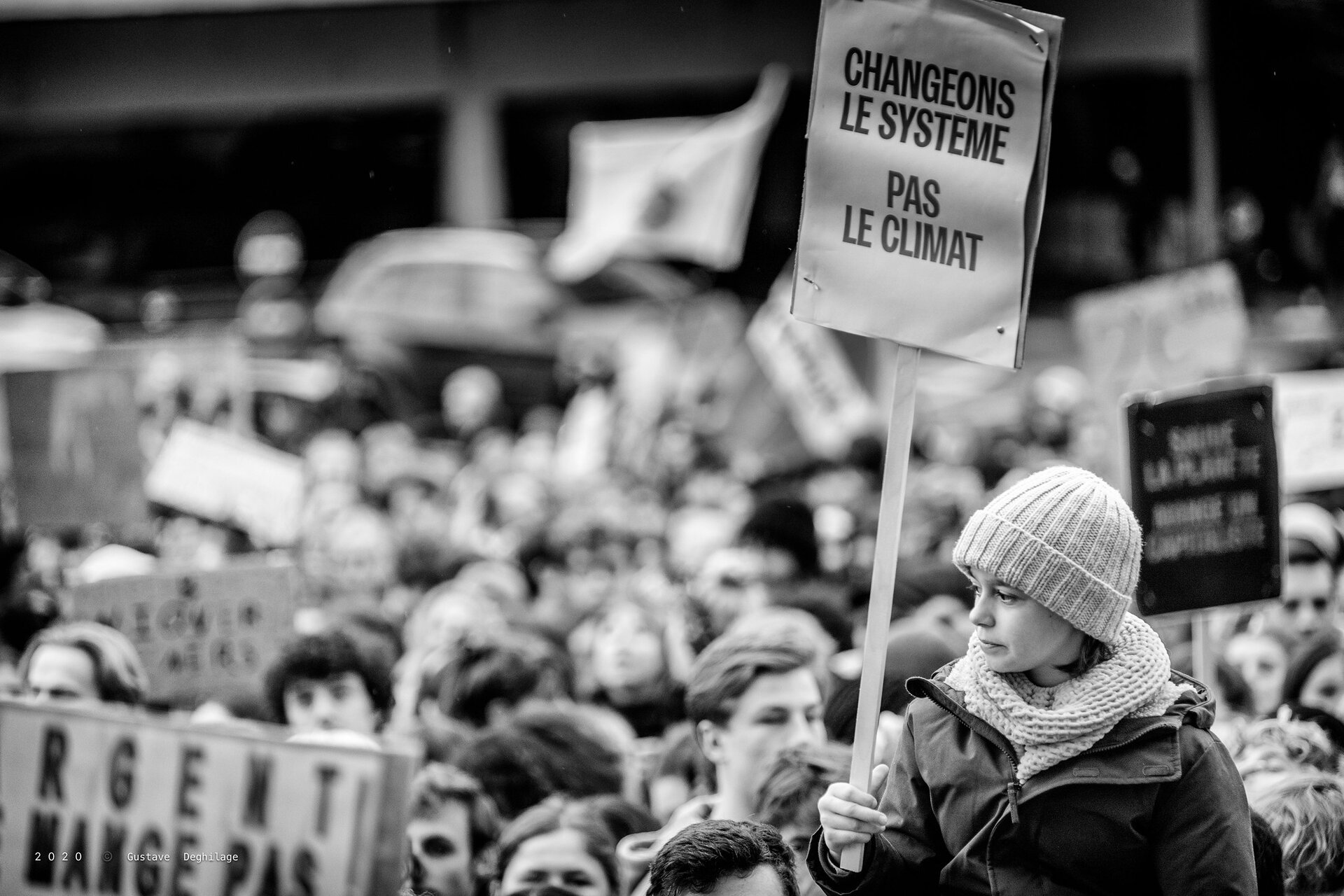 Czarno‑białe zdjęcie przedstawia tłum protestujących ludzi, ponad którymi widzimy młodą dziewczynę w białej czapce i z transparentem w ręce. Transparent zawiera napis  Changeons le systeme pas le climat