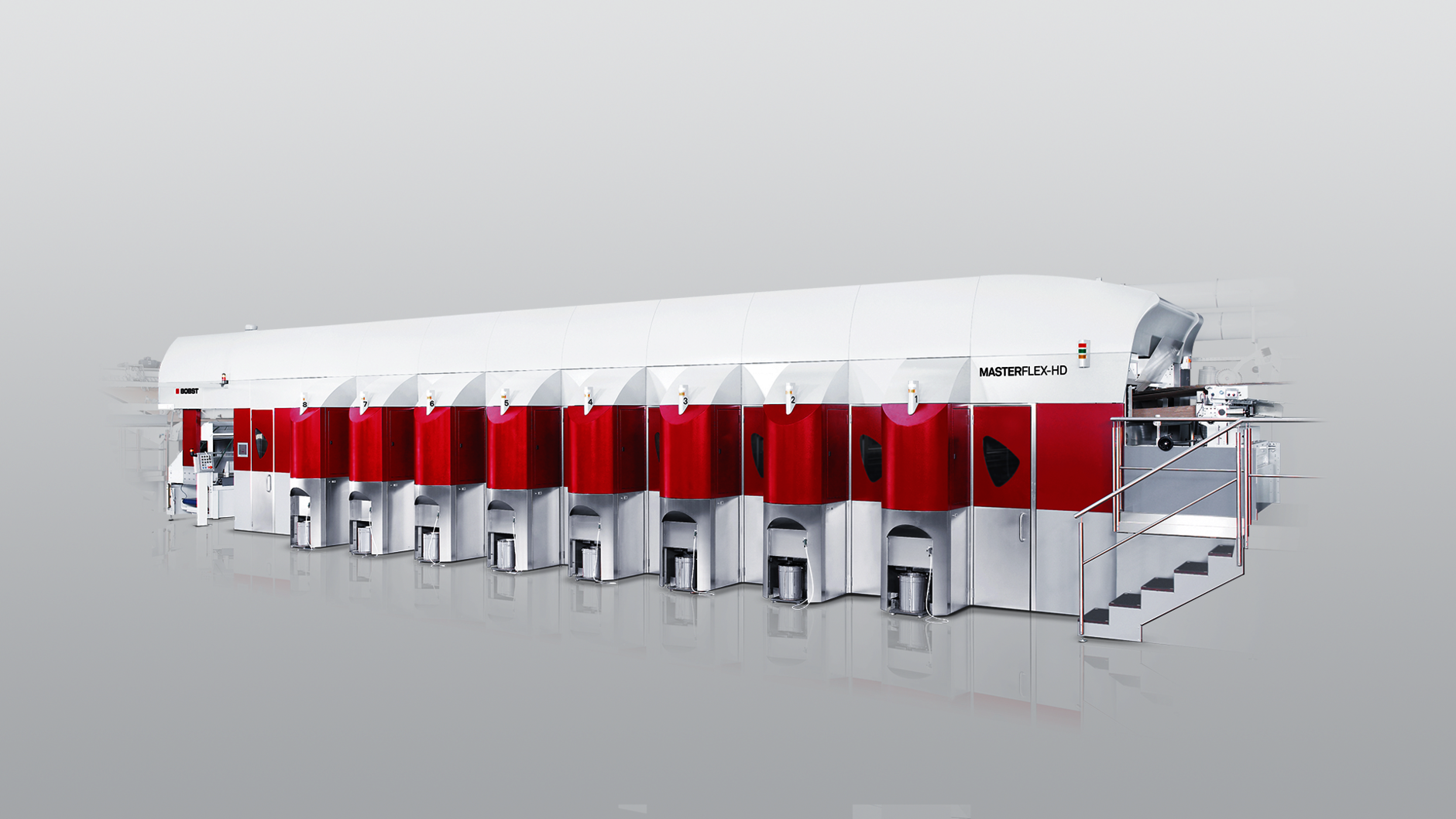 Grafika przedstawia poglądowo maszyna fleksograficzną. Maszyna ma układ szeregowy, jest zbudowana z ośmiu zespołów drukująco‑farbowych. Na przodzie maszyny widoczne schodki, które prowadzą do panelu sterującego maszyną.