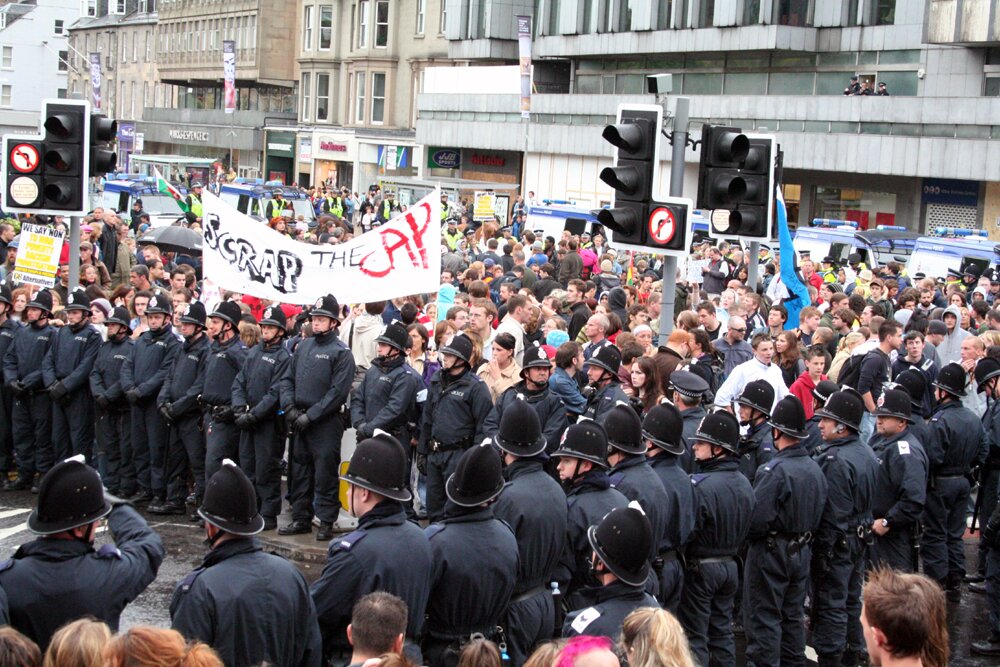 Zdjęcie przedstawia tłum ludzi w mieście, w Wielkiej Brytanii. Przed ludźmi stoi do nich ustawiony tyłem rząd policjantów. Osoby z tłumu trzymają transparent z napisem "Scrap the CAP". 