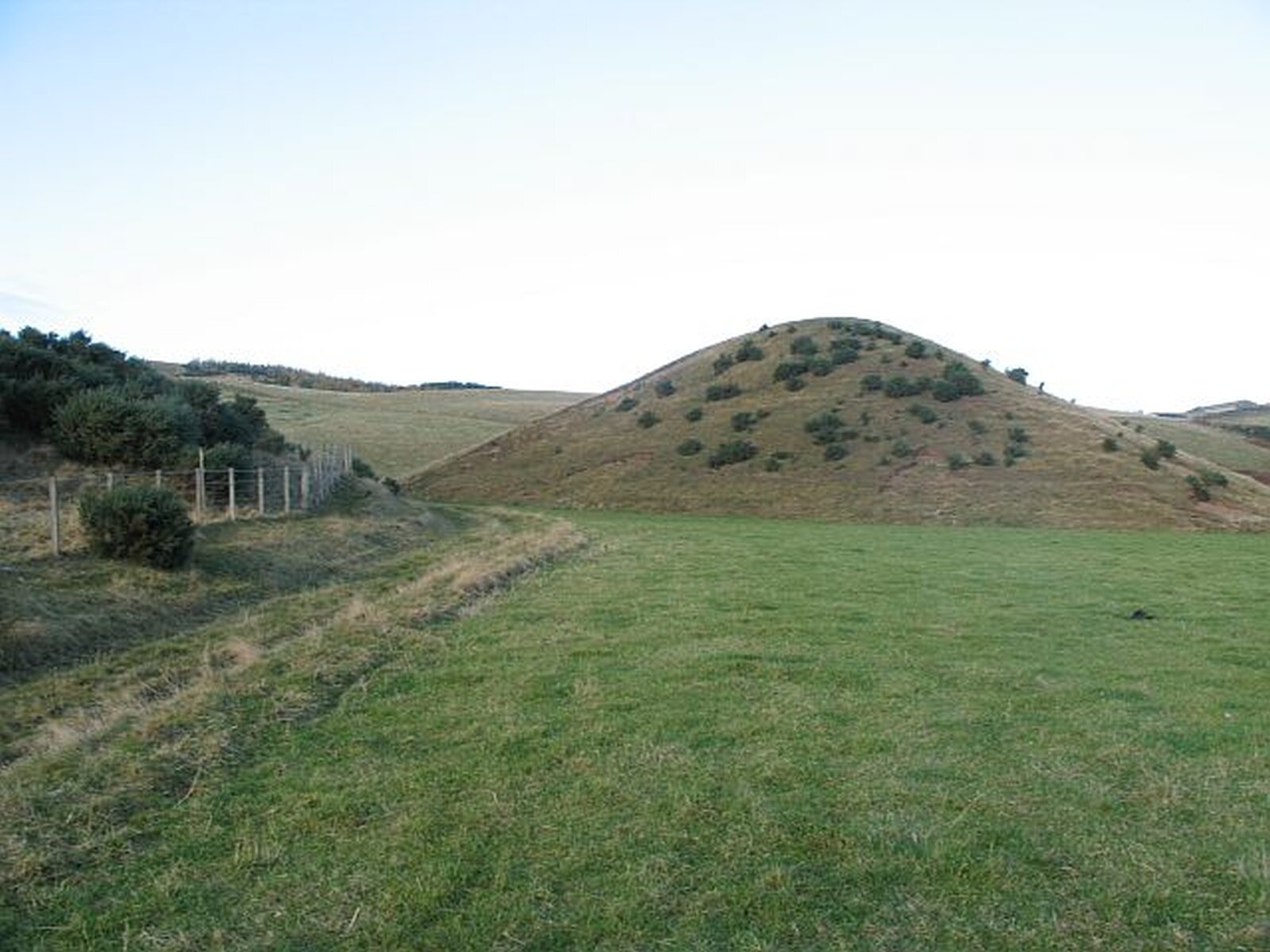 Na zdjęciu teren pokryty trawą, pośrodku niego wzniesienie o wysokości kilkunastu metrów przypominające kopiec.