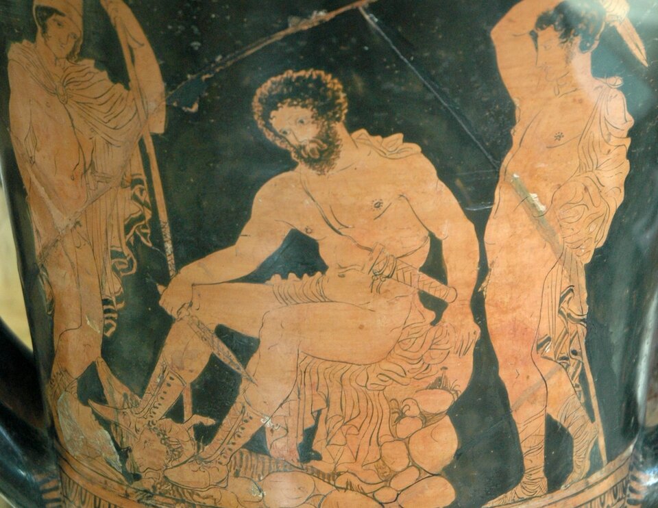 Zdjęcie przedstawia fragment starożytnej wazy czerwonofigurowej. Na środku siedzi brodaty, nagi, atletycznie zbudowany mężczyzna, który trzyma sztylet. Obok z prawej strony stoi drugi, nagi mężczyzna, atletycznie zbudowany. Jedynie na plecach ma narzuconą szatę. Trzyma uniesiony w górze sztylet. Po lewej stronie stoi mężczyzna opierający się na włóczni. Jest nagi, okryty jedynie z tyłu i na ramionach szatą.