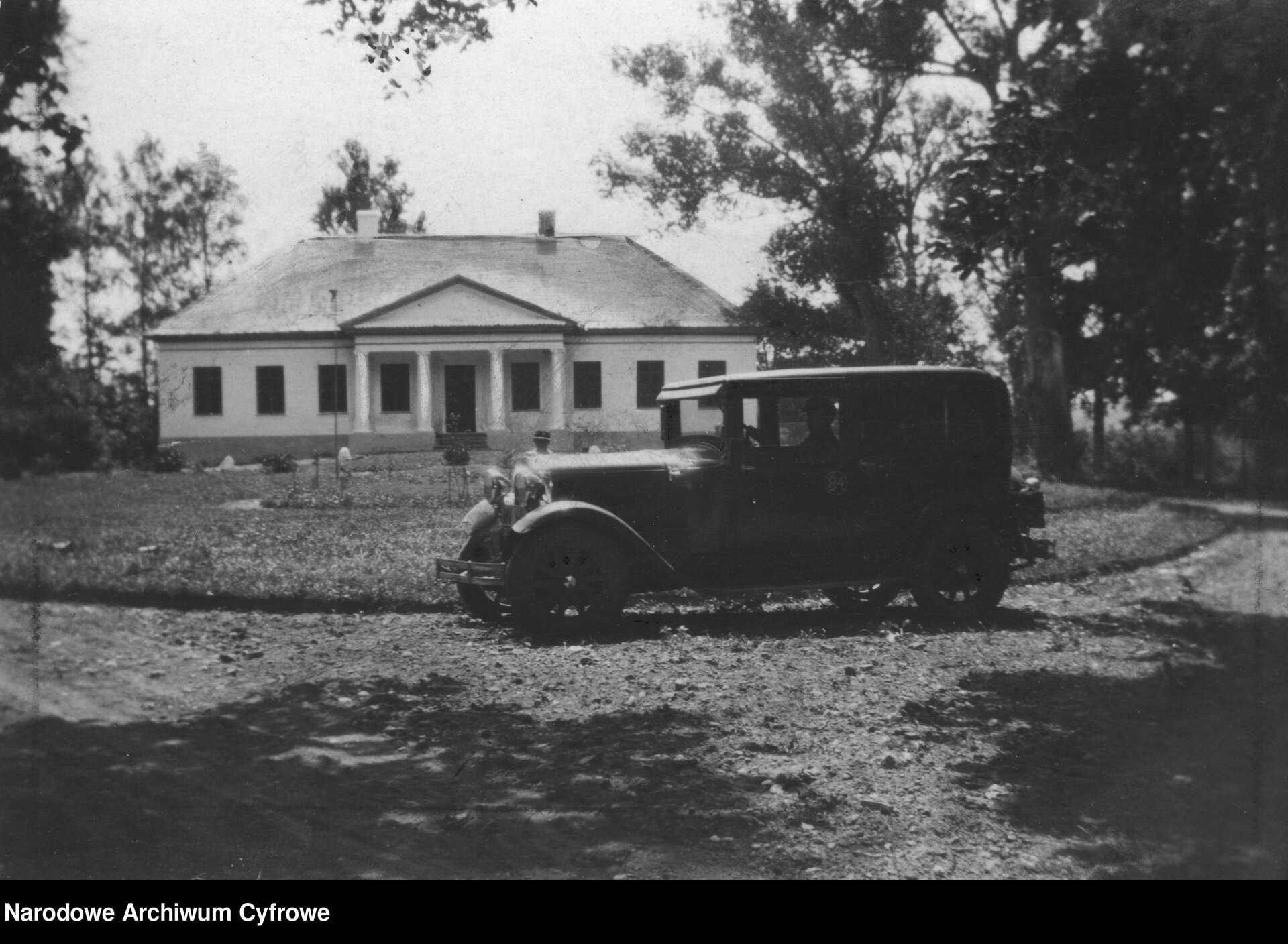 Zdjęcie przedstawia samochód osobowy w typie Forda T przed pałacykiem. Na pierwszym planie pojazd z charakterystyczną, prostokątną ramą dachu, kołami ze szprychami i zaokrąglonym nadkolem z przodu. Stoi bokiem od strony kierowcy na podjeździe do pałacu. W tle prostokątny budynek z dwuspadowym dachem i portykiem z czterema kolumnami i tympanonem po frontowej stronie budynku. Dookoła pałacu i podjazdu do niego znajdują się drzewa.