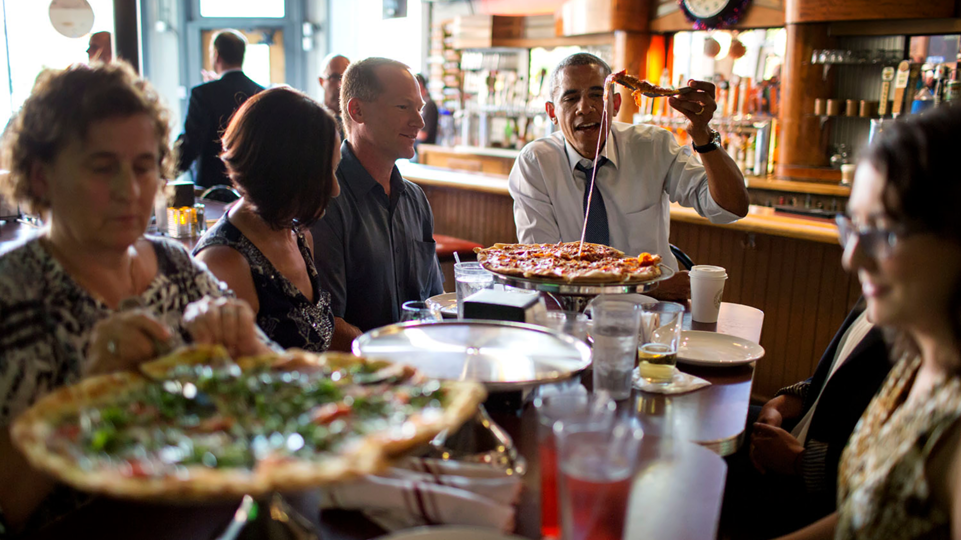 Zdjęcie przedstawia pięcioro ludzi przy stole w restauracji. Przy stole siedzą trzy kobiety i dwóch mężczyzn. Jednym z mężczyzn jest Barack Obama. Na stole znajdują się dwie pizze, talerzyki, szklanki z napojami. Barack Obama bierze w rękę kawałek pizzy, widoczny jest ciągnący się ser. W tle widoczni są inni ludzie oraz bar w restauracji.