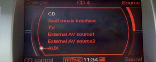 Zdjęcie przedstawia czarny prostokąt – ekran – który po prawej i lewej stronie jest otoczony przez szare i ciemno szare pola (obudowę). W czarny prostokąt są wpisane różne elementy w kolorze czerwonym, czyli napisy informujące o pomocniczej ścieżce oprogramowania aux, wpisane w spłaszczoną elipsę, w której prawym górnym rogu znajduje się pogrubiona, krótka czerwona kreska, wygięta w delikatny łuk. W czarny prostokąt wpisane są również białe elementy, czyli napis CD oraz minutnik wskazujący 11:34.