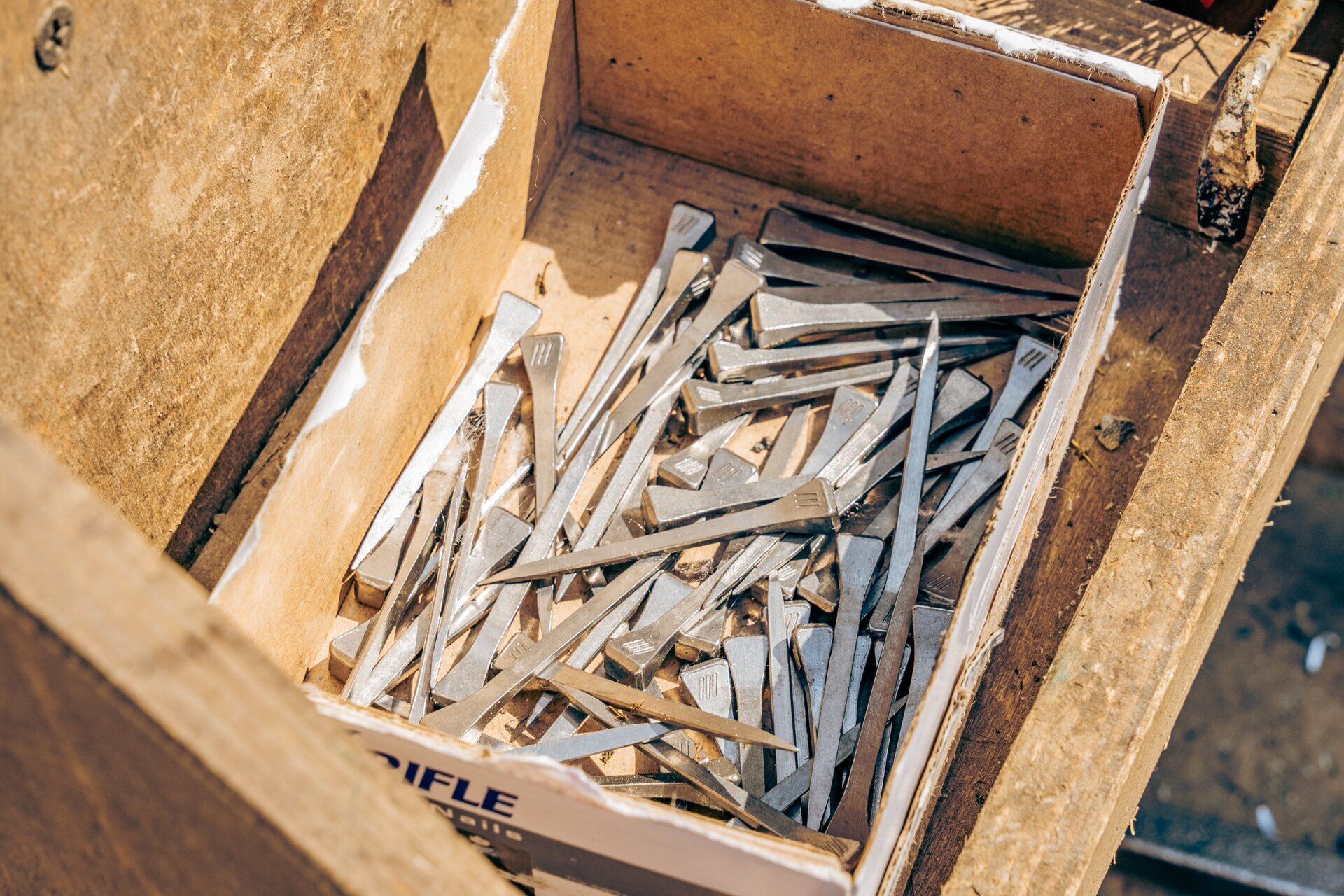 Zdjęcie przedstawia podkowiaki znajdujące się w drewnianej skrzynce. To długie metalowe elementy, które z jednej strony są ostro zakończone, a z drugiej są delikatnie poszerzone, zakończone płaską powierzchnią.