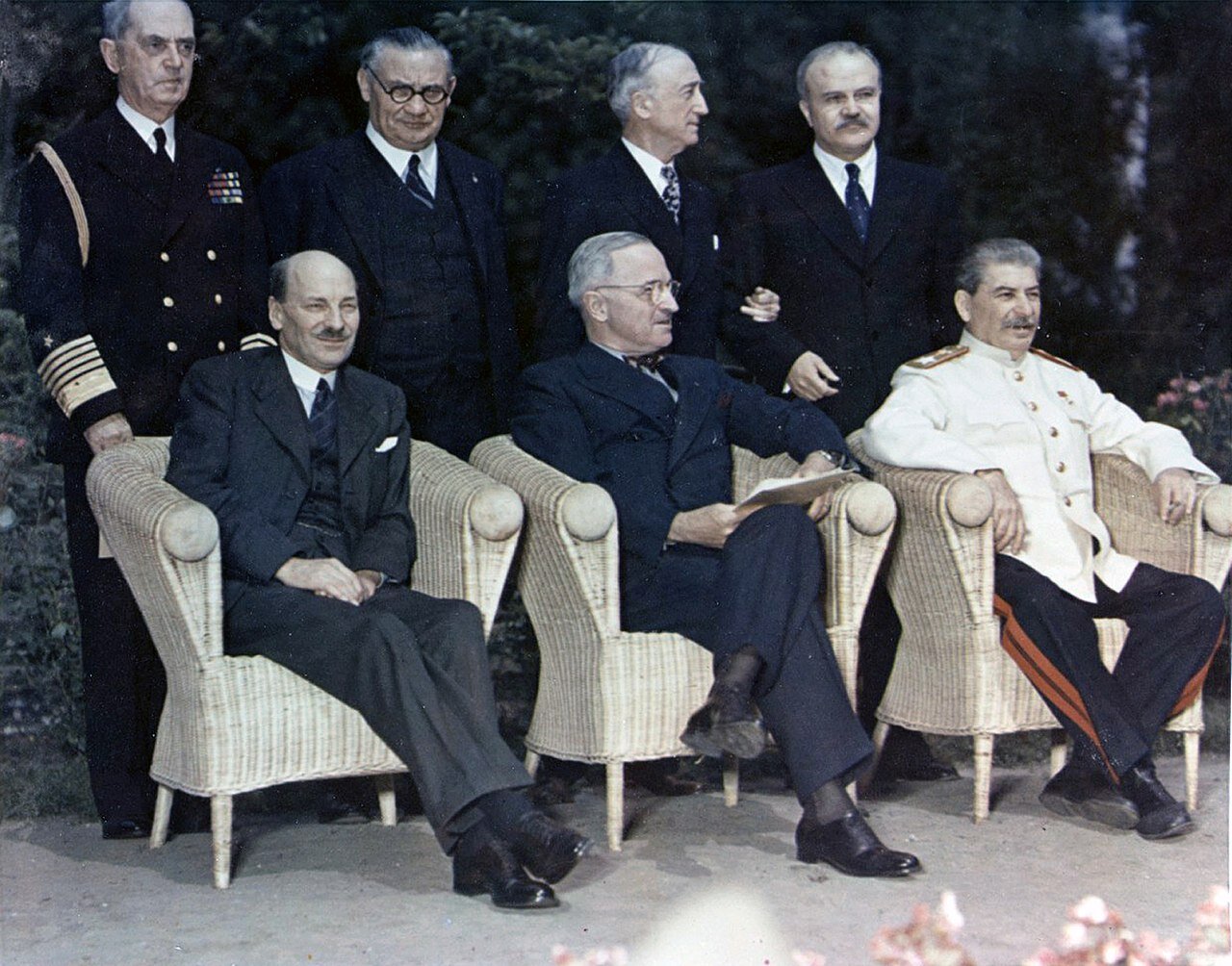 Zdjęcie przedstawia trzech mężczyzn, którzy siedzą na krzesłach. Dwaj pierwsi od lewej ubrani są w garnitury. Mężczyzna po prawej ubrany jest w jasny mundur. Za mężczyznami stoją czterej mężczyźni. Pierwszy od lewej ubrany jest w mundur, pozostali w garnitury.