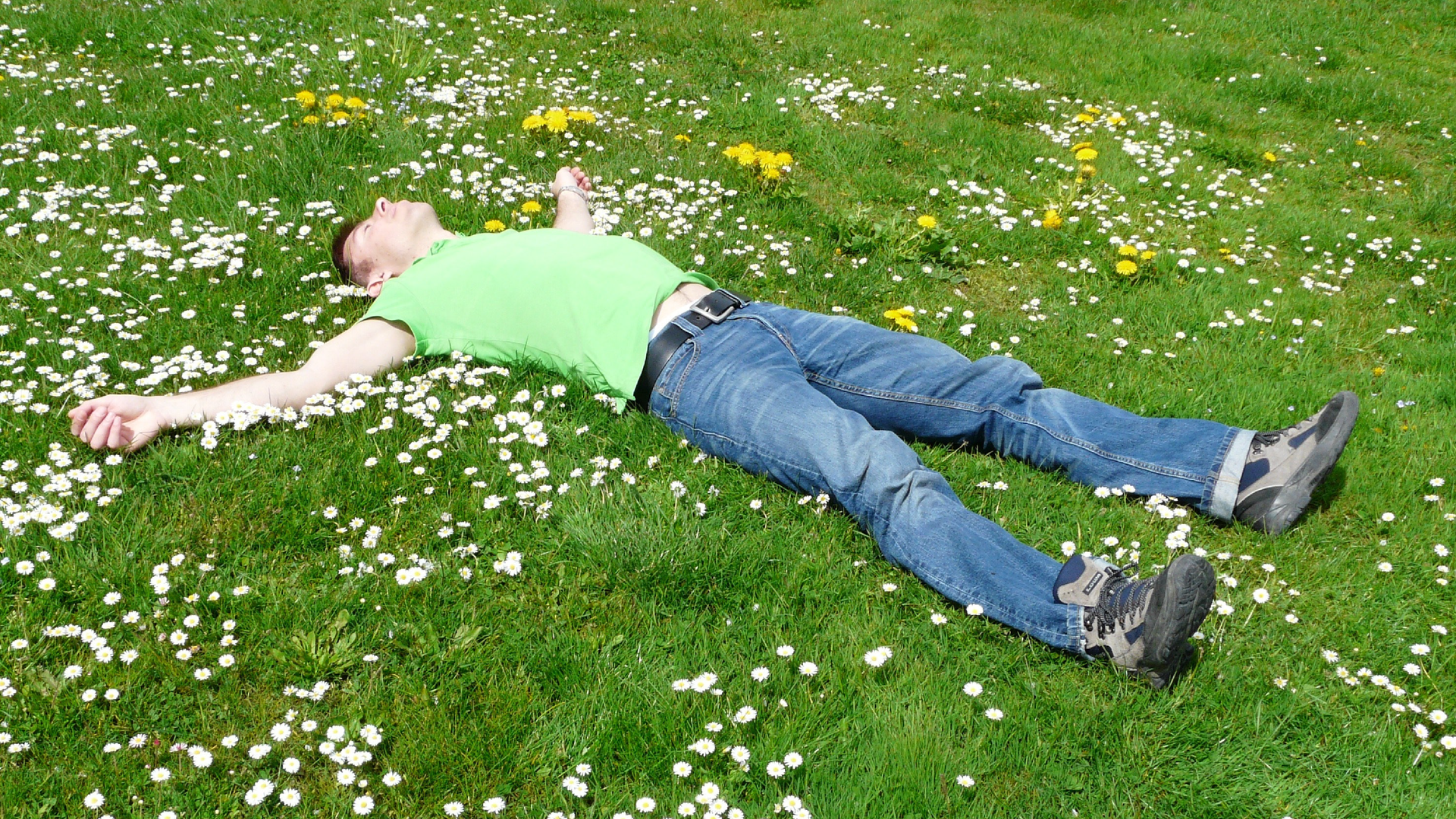 Na zdjęciu znajduje się mężczyzna, który leży na wznak na łące pośród kwiatów i zielonej trawy. Mężczyzna ma zamknięte oczy i delikatnie się uśmiecha.