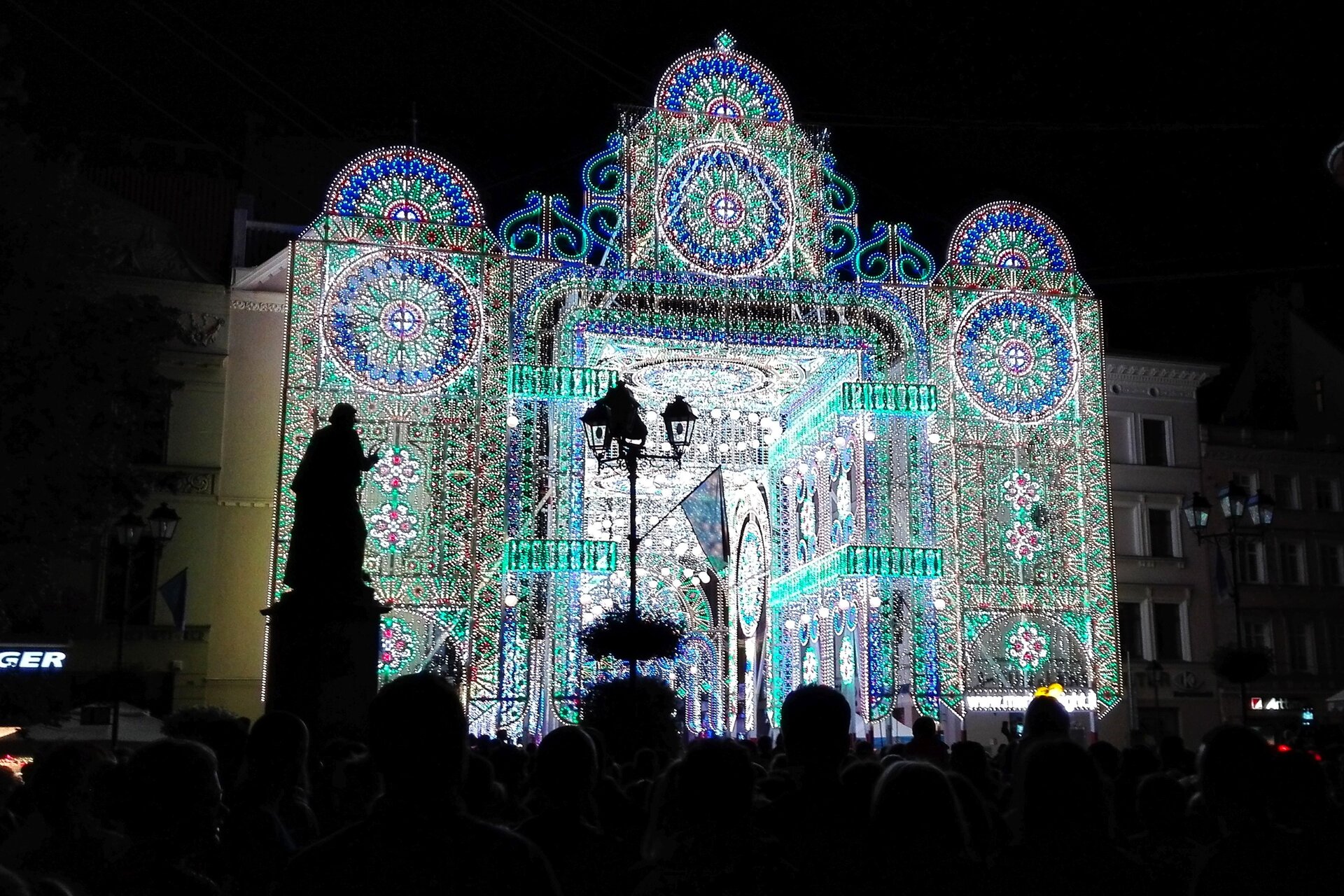 Na zdjęciu stoi budynek, na którym wyświetlony został obraz z geometrycznymi wzorami. Na budowli wyświetlają się następujące kolory: biały, zielony, niebieski, fioletowy. Przed budynkiem stoi tłum ludzi, latarnia z flagą i posąg. Tło stanowi zabudowa miejska i czarne nocne niebo. Fotografia jest ciemna, jedynie budynek i jego najbliższe otoczenie jest oświetlone. 