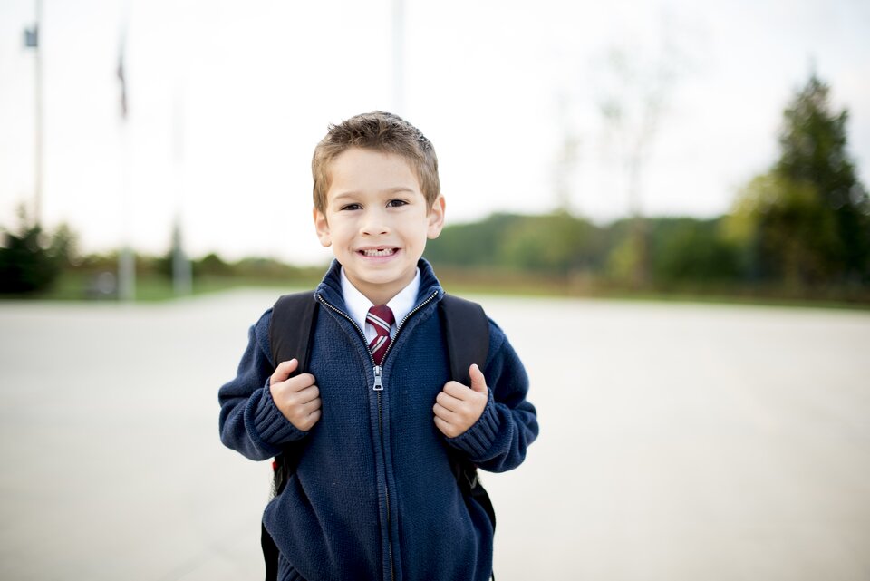 Zdjęcie ukazujące chłopca w wieku szkolnym. Ubrany jest w niebieski, zapinany na zamek sweter. Pod swetrem ma koszulę z kołnierzykiem i czerwony krawat. Na plecach chłopiec ma plecak.