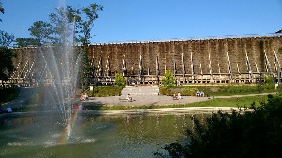 Zdjęcie przedstawia tężnie solankowe. Na pierwszym planie znajduje się fontanna. Za nią jest plac z ławkami, na których siedzą ludzie. Dalej znajdują się wysokie tężnie. Podtrzymywane są drewnianą konstrukcją.