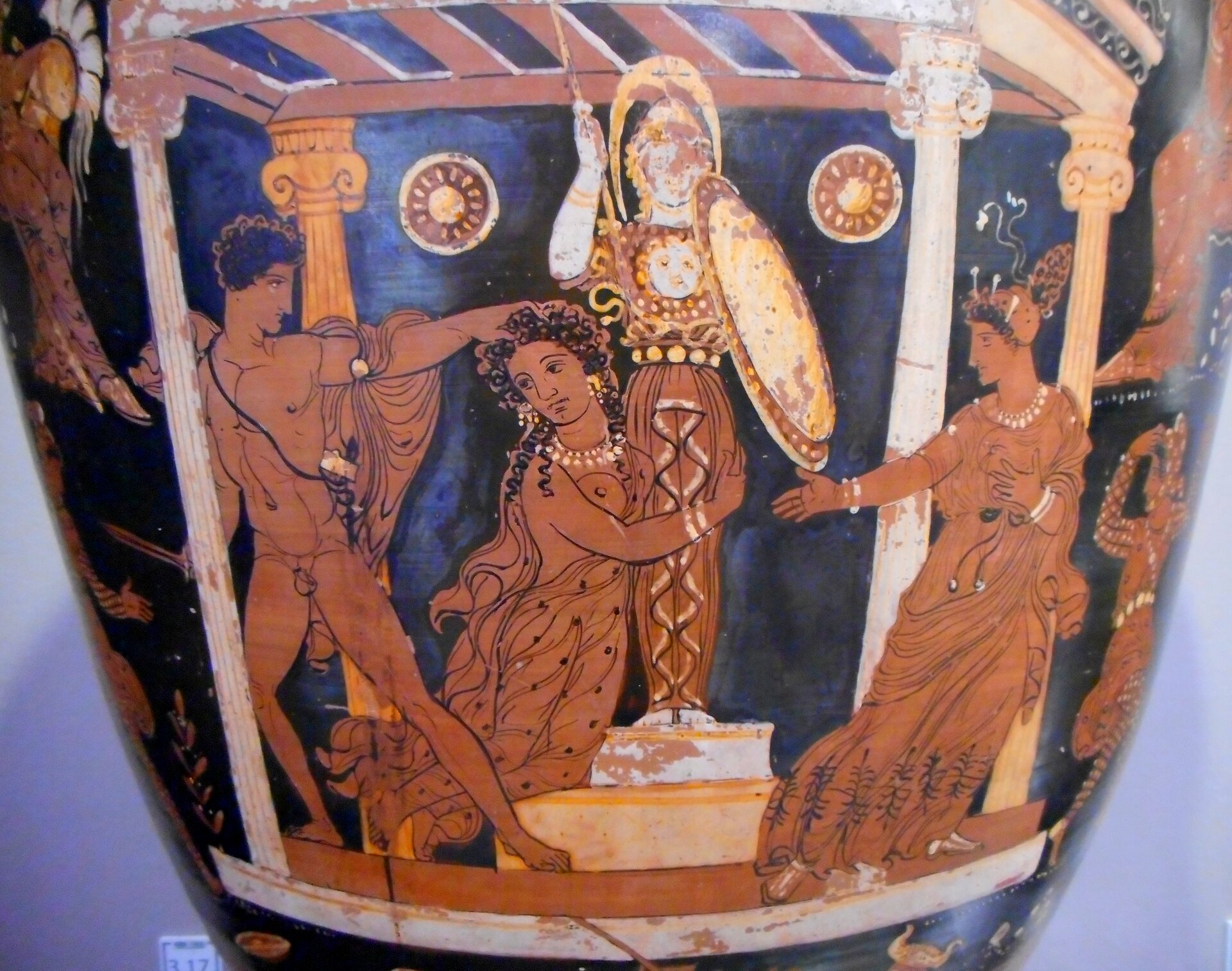 Fotografia nieznanego autora przedstawia przykład malarstwa czerwonofigurowego na ciemnej wazie. Dzieło prezentuje trzy postacie. Po lewej stronie znajduje się nagi mężczyzna, który jedną ręką przytrzymuje nóż, a drugą robi zamach aby zadać cios. Znajdująca się w centrum kobieta ma długie ciemne włosy, jest ubrana w długą suknię, spogląda na agresora, przytula się do figury bogini Ateny. Druga kobieta znajduje się z prawej strony naczynia. Jest przedstawiona jako kobieta w średnim wieku. Ma krótkie czarne włosy, przykryte czapką. Ubrana jest w długą suknię. Na szyi oraz dłoniach widoczne są korale. Jedną dłoń trzyma na piersi, drugą wyciąga do zaatakowanej kobiety.