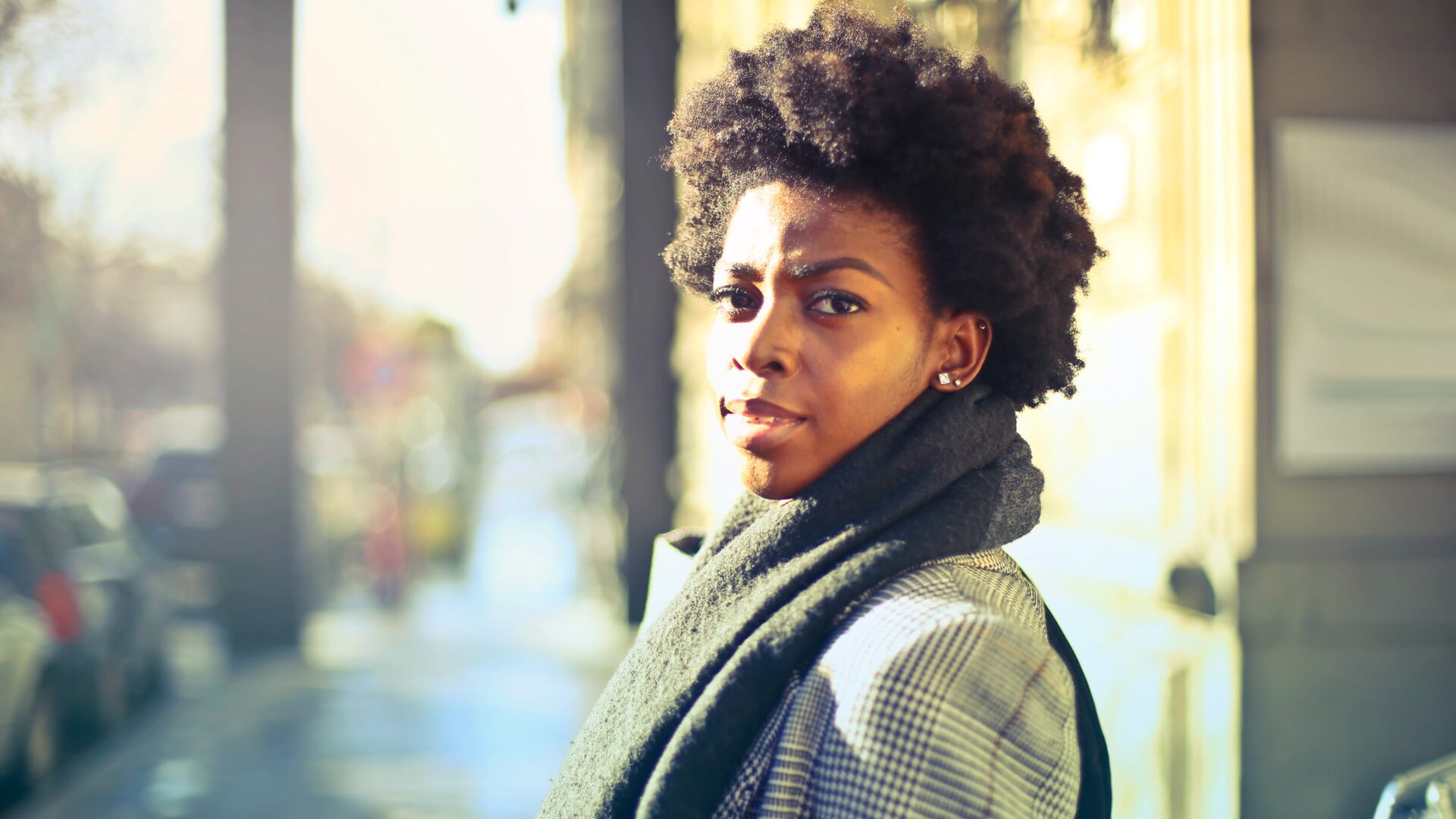 Zdjęcie przedstawia ciemnoskórą, młodą kobietę, stojącą na chodniku. Patrzy w obiektyw aparatu. Ma krótkie, bardzo gęste ciemne włosy, pełne usta, ciemne oczy, długie brwi. Ubrana jest elegancko - w kraciastą marynarkę, na szyi ma szal. W tle przy chodniku po lewej stronie stoją zaparkowane samochody, po prawej stronie chodnika jest budynek.  