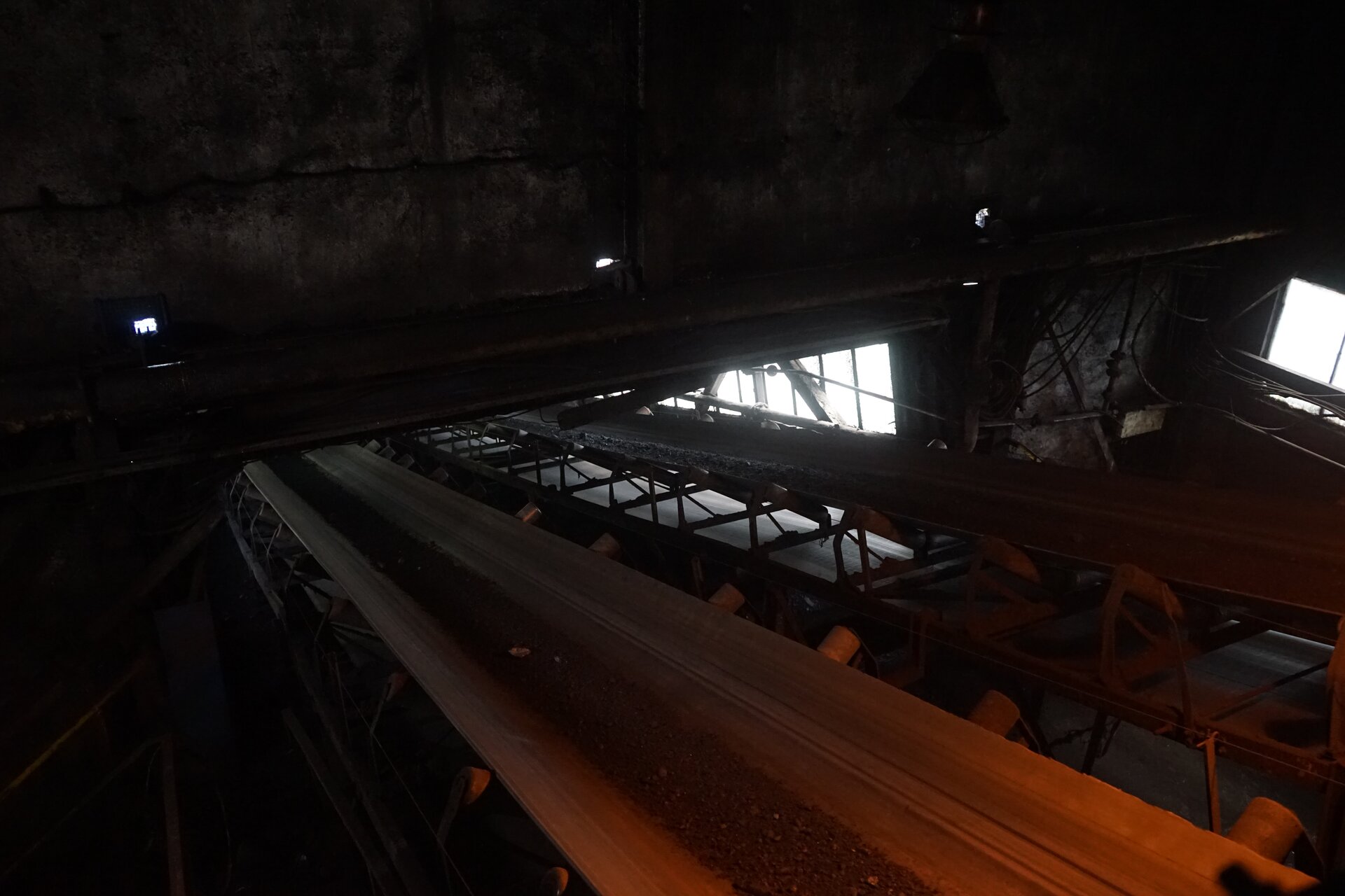 Na zdjęciu widać fragment dwóch równoległych przenośników taśmowych czyli urządzeń wykorzystywanych do transportu materiału przy użyciu taśmy na dalsze odległości, które znajdują się wewnątrz obiektu budowlanego.Podstawą przenośników taśmowych jest widoczna u dołu zdjęcia stalowa konstrukcja kratowa czyli trasa. Konstrukcja ta zbudowana jest powtarzalnych członów modułów których podstawą są ceowniki. Ceownik jest to wyrób hutniczy, walcowany lub gięty z metalu. Wyroby te są w postaci prostych odcinków. Przekrój poprzeczny czyli profil ceownika przypomina literę C. Pojedynczy boczny element trasy jest w kształcie rdzawego prostokąta.Z boku konstrukcji czyli na boku trasy znajdują się metalowe elementy w kształcie ceowników, które biegną pionowo w górę. Końcówki tych elementów są zakrzywione w kierunku taśmy czyli w kierunku środka przenośnika. Są to kozły górne. Kozły górne są rozmieszczone po oby stronach trasy. Pomiędzy kozłem górnym a wspornikiem bocznym rolki dźwigara znajduje się krążnik górny który na zdjęciu jest w kształcie czerwonego walca. Na krążniku osadzona jest szara taśma w kształcie odwróconej litery C, która biegnie przez całą trasę przenośnika. Na taśmach obu przenośników znajdują się drobne bryłki węgla. W dalszych fragmentach przenośnika widać z boku konstrukcji trasy odchodzące pionowo w dół krótkie stalowe elementy w kształcie ceowników. Są to kozły dolne. Na walcowatych elementach umieszczonych po środku kozła dolnego osadzony jest krążnik dolny. Czerwone krążniki dolne są również w kształcie walców. Na krążnikach dolnych znajduje się taśma, która ulega zawróceniu.Oba widoczne na zdjęciu przenośniki są nieznacznie odchylone od poziomu co wskazuje na transport materiału pod kątem. Po lewej stronie przenośnika znajdującego się po lewej stronnie znajduje się wąskie przejście. Po prawej stronie przenośnika znajdującego się po prawej stronie widać podłużne okna. Pomieszczenie przemysłowe przez które biegną przenośniki zbudowane jest z betonu i charakteryzuje się niską wysokością. 