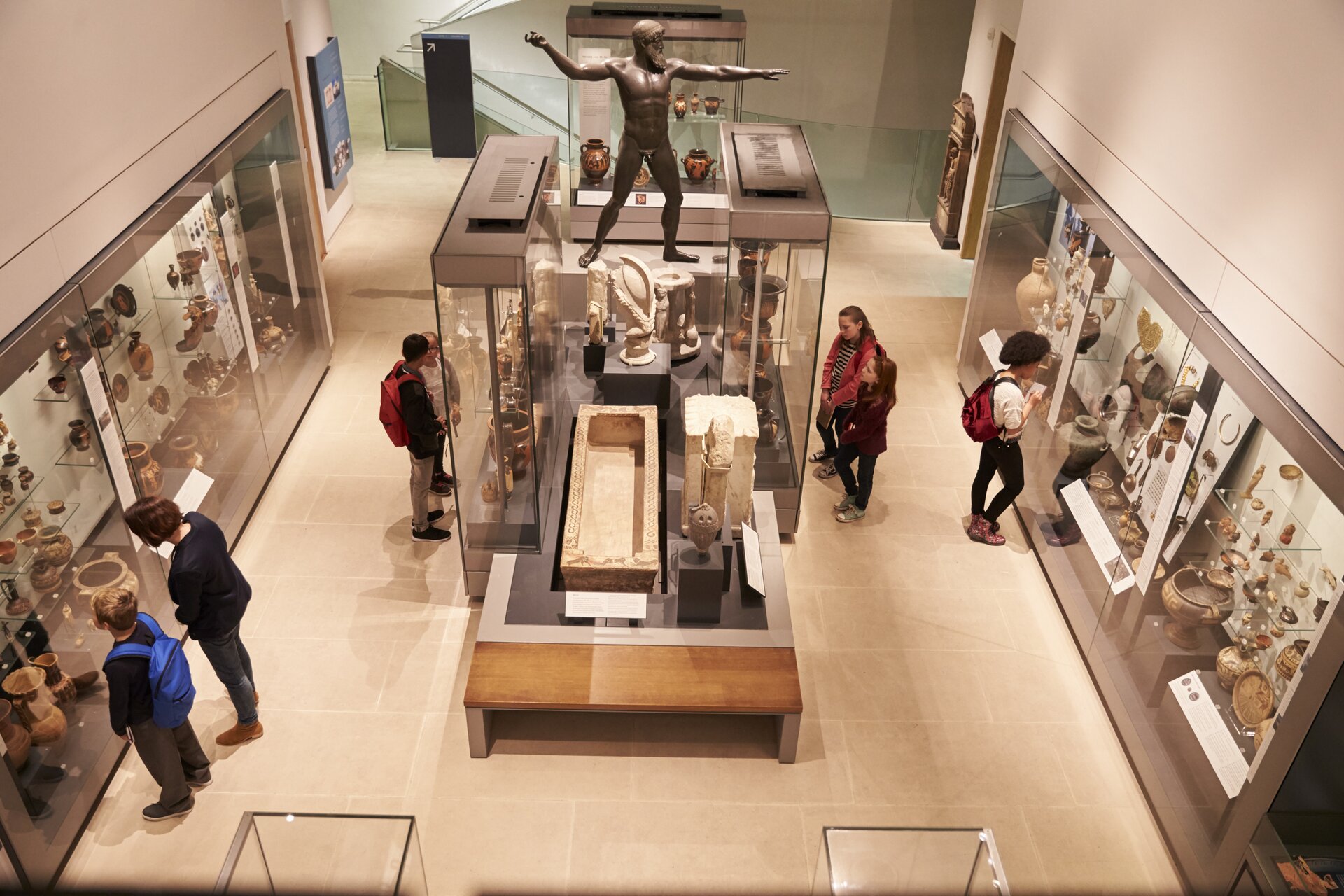 ilustracja przedstawia wnętrze muzeum. Na ścianach po lewej i prawej stronie rozmieszczone są oszklone gabloty z eksponatami sztuki starożytnej. W środku sali znajduje się duża przeszklona gablota a obok niej ekspozycja prezentowana na drewnianym podeście w kształcie stołu. W muzealnej sali widoczne są osoby oglądające prezentowane zbiory.