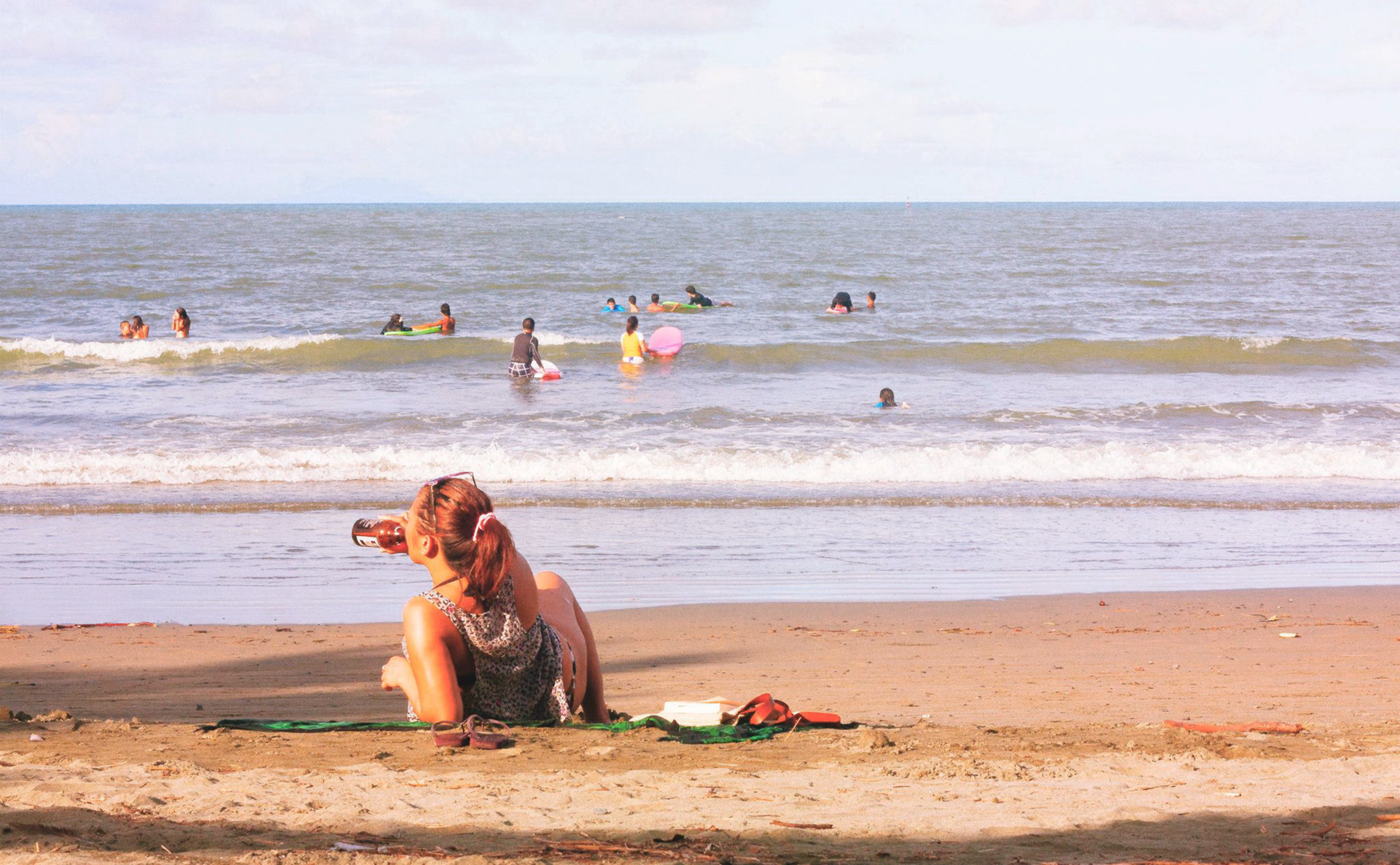Kolorowe prostokątne zdjęcie przedstawia słoneczny letni dzień na plaży. Na pierwszym planie młoda kobieta leży na kocu, na piasku. Wsparta na prawym łokciu. W drugiej ręce trzyma butelkę z piwem. Pije piwo z butelki. W głębi morze. Małe fale. Kilkanaście osób pływa w morzu.