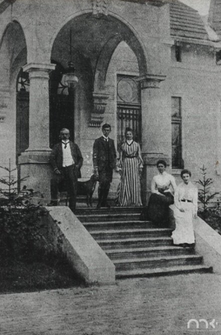 Zdjęcie przedstawia zbliżenie wejścia do pałacu. Ukazuje pięć osób. Dwie kobiety w sile wieku siedzą na niskim murku znajdującego się po bokach schodów. Są to szczupłe osoby. Mają włosy upięte z tyłu głowy. Są ubrane w jasne bluzki i długie spódnice. Na szczycie schodów stoją trzy osoby i pies. Po lewej stronie dojrzały mężczyzna opiera prawą nogę o mur. Jest ubrany w koszulę, marynarkę i spodnie. Lewą rękę trzyma w kieszeni spodni. Pośrodku stoi młody mężczyzna. Przy jego prawym boku stoi pies. Po prawej stronie stoi kobieta ubrana w długą suknię w pionowe paski.