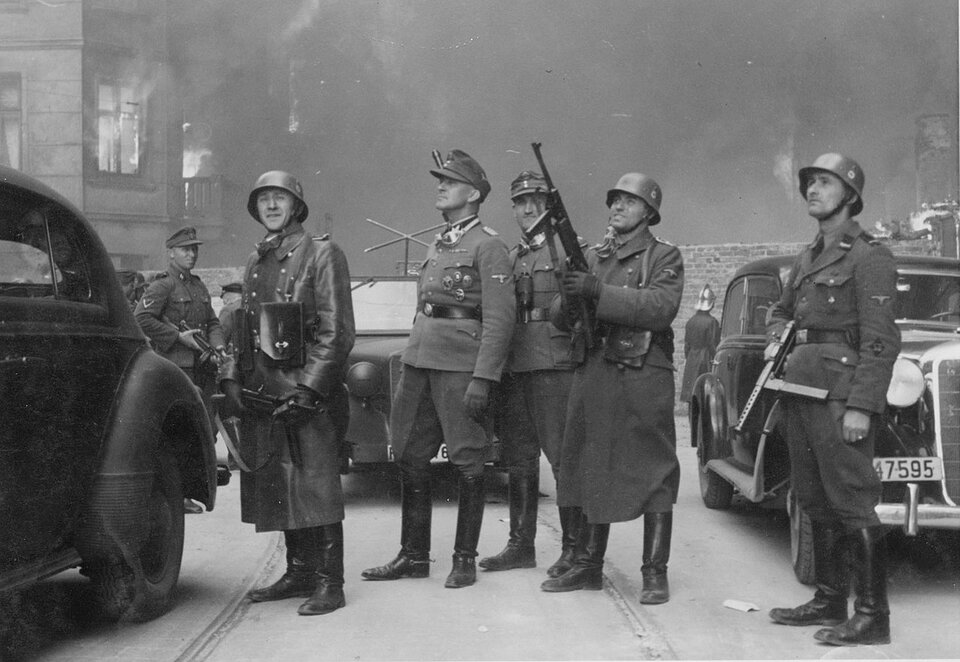 Zdjęcie przedstawia grupę żołnierzy niemieckich. Na pierwszym planie widać pięciu żołnierzy. Dwóch z nich jest w mundurach oficerskich. Trzech to zwykli żołnierze. Wszyscy są uzbrojeni. Wokół nich stoją trzy samochody osobowe. W pobliżu widać kolejnych żołnierzy. Na drugim planie widać płonącą kamienicę. W powietrzu unosi się gęsty dym.