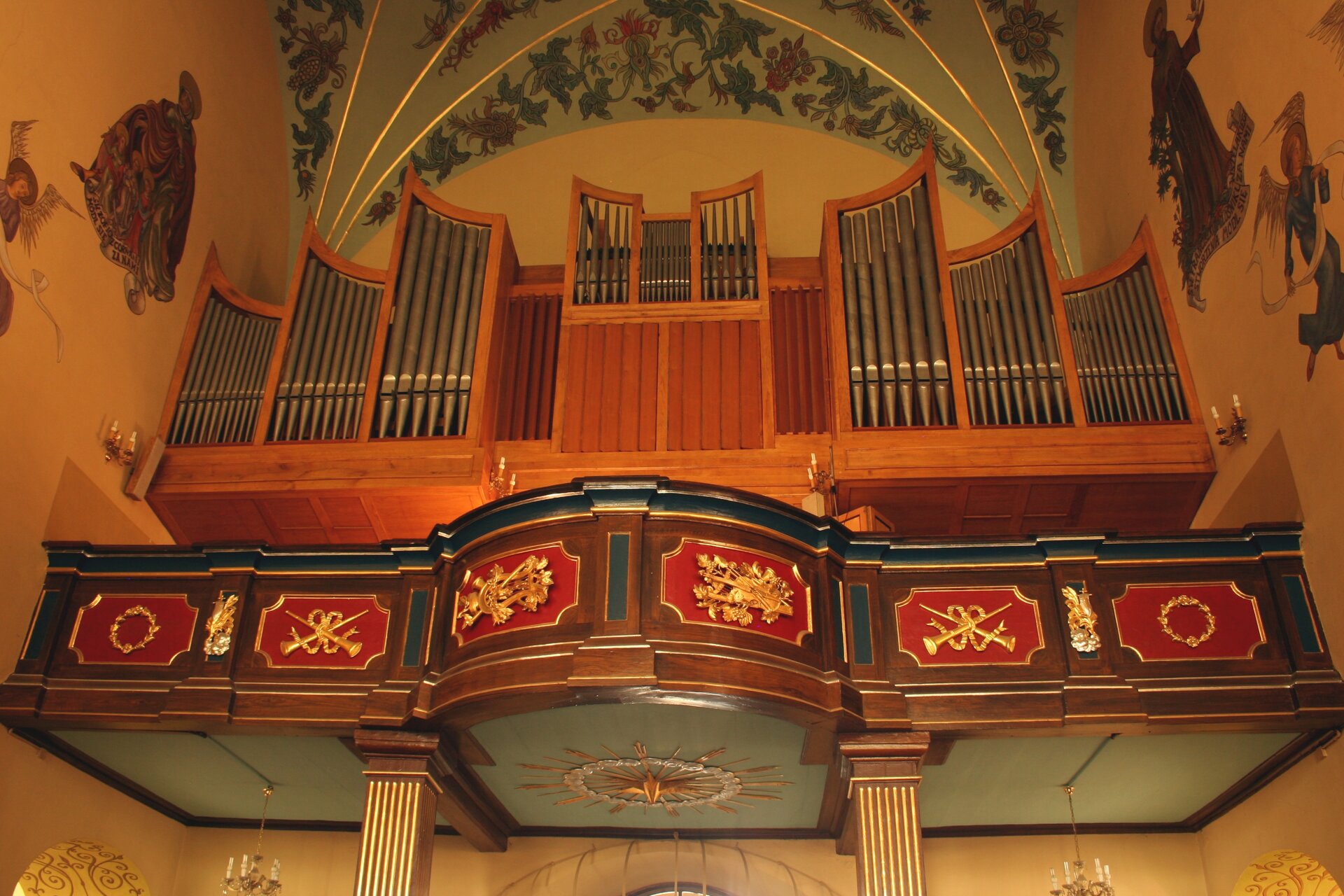Obraz przedstawia organy na zdobionym balkonie. organy wykonane są z drewna, są symetryczne.  Po lewej i prawej stronie organów znajdują się po cztery sekcje piszczałek, dodatkowo pomiędzy nimi znajduje się jedna wspólna sekcja. W pierwszej sekcji znajduje się 11 piszczałek, w drugiej 8 piszczałek, w trzeciej 6 piszczałek, w czwartej 5 piszczałek, a w piątej wspólnej sekcji znajduje się 9 piszczałek. Na sklepieniu kościoła namalowane są kwiaty, a na ścianach widnieją obrazy świętych osób oraz herubinów. Balkon wykonany jest z ciemnego drewna. Posiada on wycięcia pomalowane na czerwono, ramki są pozłacane. W środku każdego wcięcia widnieją pozłacane przedmioty. W pierwszej sekcji wieniec laurowy. W drugiej sekcji dwa długie proste, skrzyżowane rogi związane ze sobą wstążką. W trzeciej sekcji widać harfę, róg oraz liście. Prawa strona jest lustrzanym odbiciem lewej strony, z tym że pierwsza sekcja posiada skrzypce, dużą harfę, róg oraz winogrona. Pod balkonem wisi pozłacany ornament na kształt słońca otoczony szklanymi dyskami. Balkon umocowany jest do podłoża solidnymi drewnianymi filarami. Pod balkonem widać dwa zwisające żyrandole.