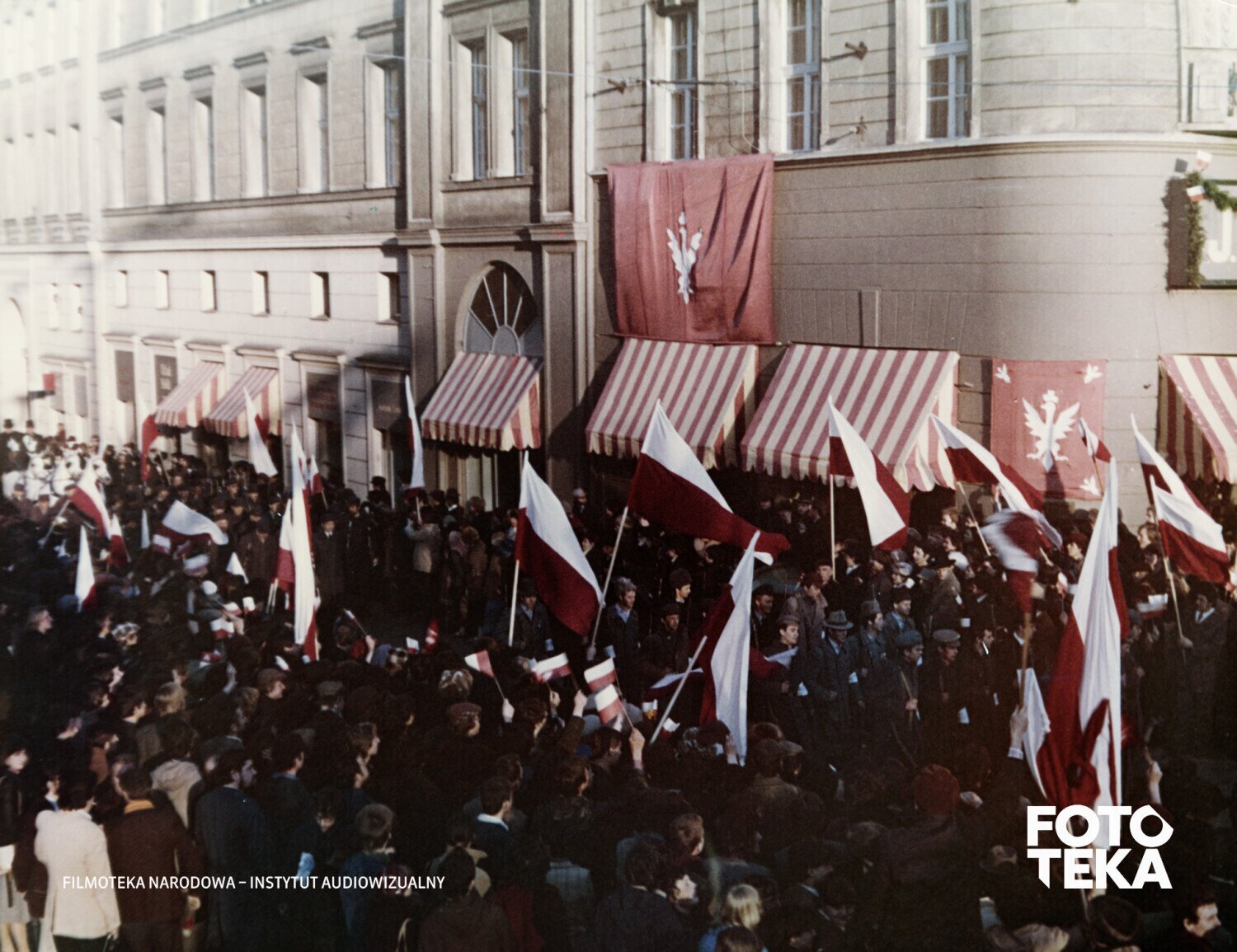 Kolorowa fotografia przedstawia ulicę miasta wypełnioną tłumem trzymającym polskie flagi. 