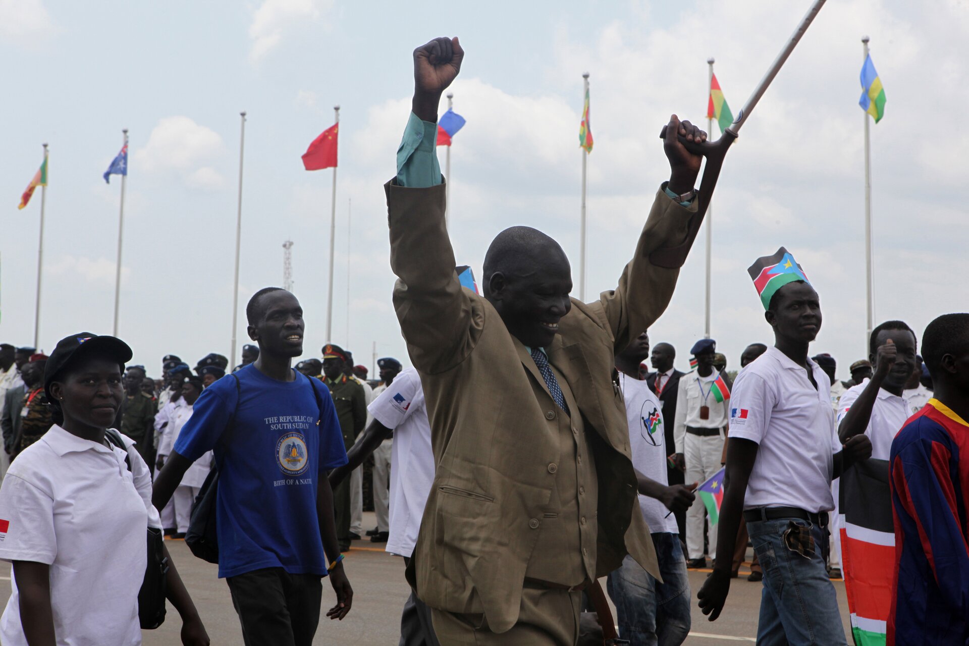 Zdjęcie przedstawia tłum czarnoskórych ludzi. Mężczyzna na pierwszym planie ma uniesione do góry ręce w geście radości. Po prawej stronie idących ludzi stoją w rzędzie wyższej wojskowi odświętnie ubrani. Za nimi powiewają flagi różnych państw.
