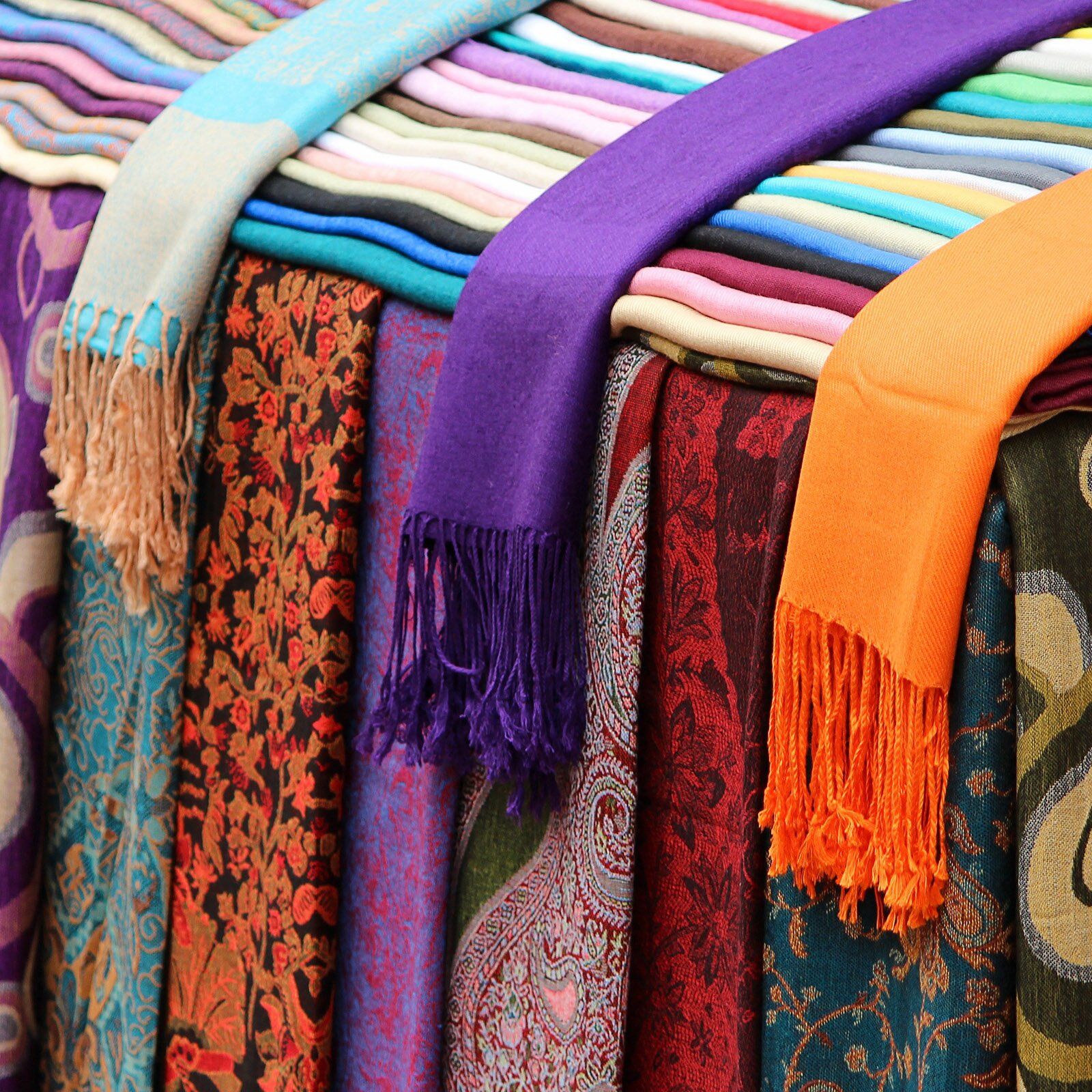  Zdjęcie przedstawia ułożone szale i chusty. Wzorzyste szale zwisają i zachodzą na siebie, natomiast szale w jednolitych kolorach są poukładane w harmonijkę. Na nich leżą trzy szale z frędzlami. Mają kolor pomarańczowy, fioletowy i beżowo‑niebieski. 