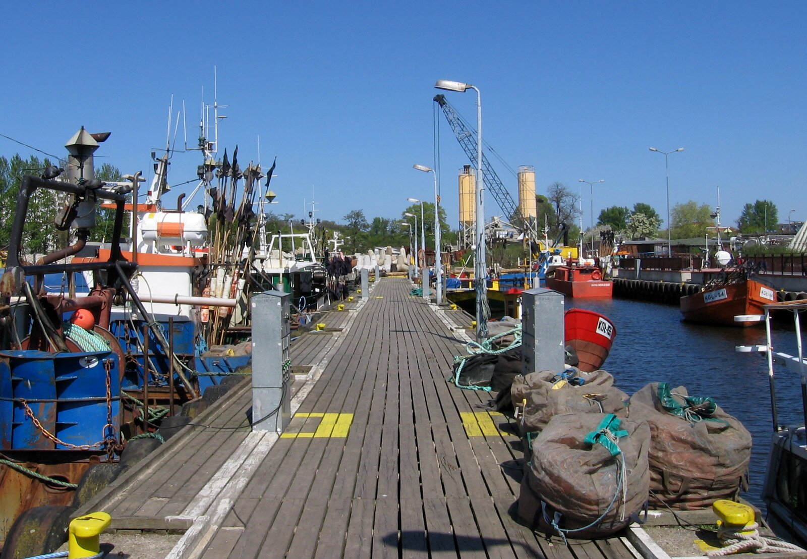 Zdjęcie ukazuje drewniany pomost, przy którym znajdują się różne maszyny i łodzie. Na pomoście znajdują się lampy i przedmioty związane z rybactwem. W dalszej części ilustracji widać zalesiony teren.