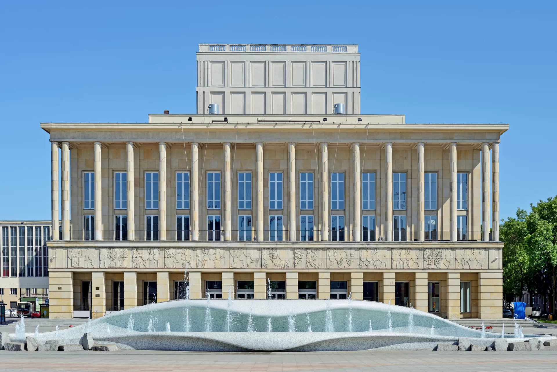 Fotografia przedstawia Teatr Wielki w Łodzi. Jest to budynek w kształcie prostokąta. Fronton o długości 50 metrów składa się z podwójnego ażuru i loggii. Główne wejście znajduje się w podcieni. Loggia opiera się o przestrzeń foyer. Górna część budynku, z wielkimi oknami, ozdobiona jest okrągłymi, wąskimi kolumnami. Dolna część krótszymi, kwadratowymi kolumnami. Pomiędzy górną, a dolną częścią znajduje się pas rzeźbiarskiego fryzu (ok. 5 m nad terenem), na którym przedstawione zostały antyczne wzory (pegazy, konie, ludzkie postacie, elementy dekoracyjne). Przed budynkiem znajduje się asymetryczna fontanna w kształcie dwóch morskich fal. Teatr Wielki jest drugim pod względem wielkości gmachem operowym w Polsce. Obecnie, widownia liczy 1074 miejsc.