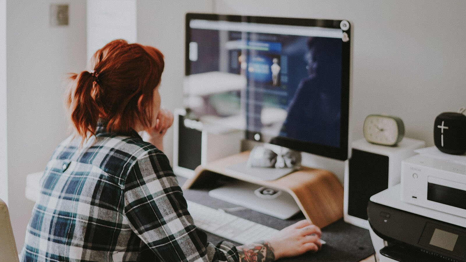 Zdjęcie przedstawia kobietę siedzącą tyłem do nas, przy biurku, przed dużym monitorem. Kobieta w prawej ręce trzyma mysz komputerową, a lewą podtrzymuje podbródek. Na biurku przed nią leży klawiatura. Obok niej po prawej stronie stoi drukarka, głośnik, a na nim zegarek.