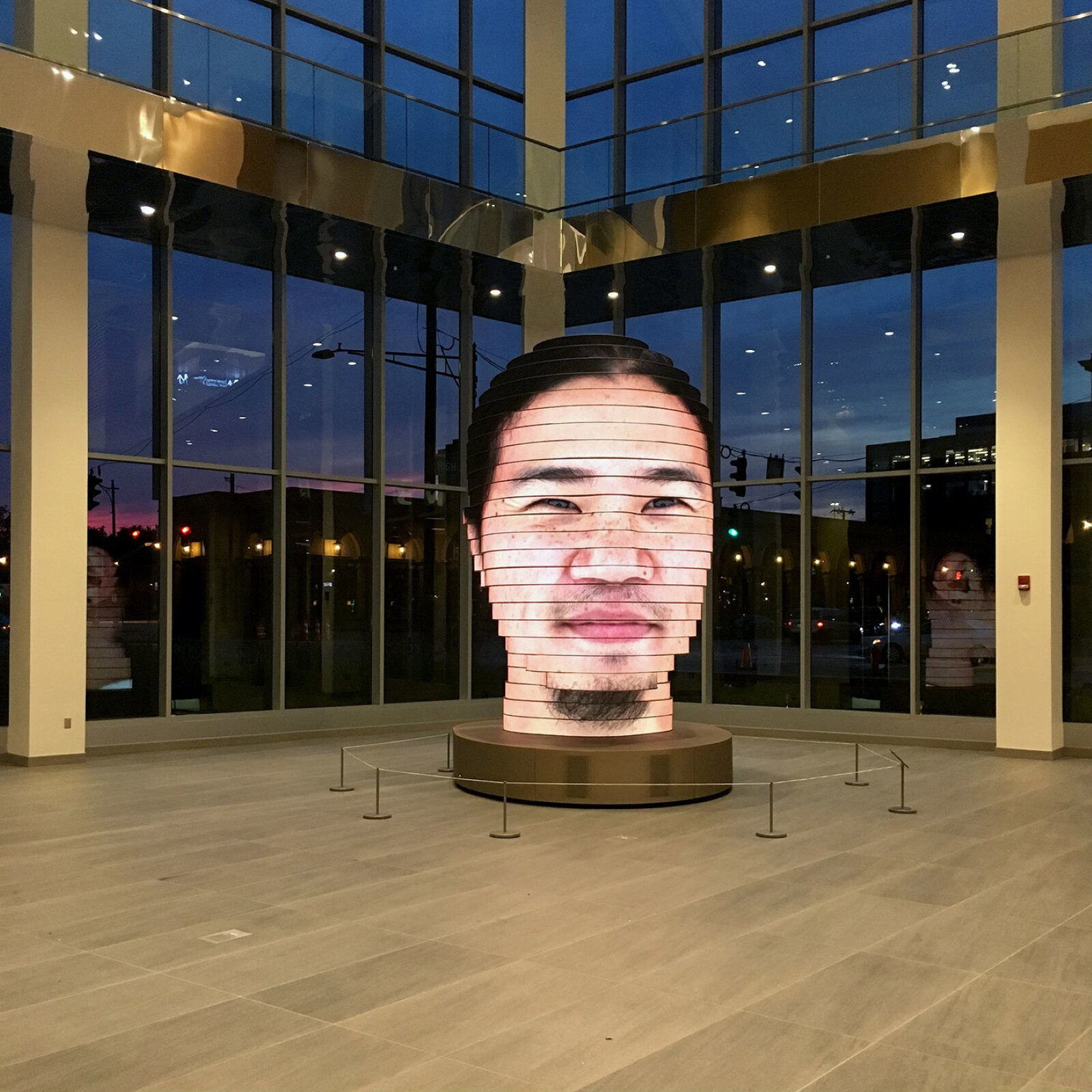 djęcie przedstawia interaktywną rzeźbę głowy. Rzeźba składa się z podłużnych paneli LED, ułożonych w poprzek, jeden na drugim. Na tych panelach wyświetlany jest obraz, który tworzy ludzką twarz. Przedstawiony został mężczyzna z piegami na twarzy, dużym nosem, małymi, przymrużonymi oczami. Mężczyzna ma zaznaczone zmarszczki wokół oczu. Ma niewielką brodę oraz wąsy. Na głowie ma ciemne włosy. Rzeźba oddaje nawet światło, które odbija się od twarzy, dzięki czemu jest bardzo realistyczna. Stoi na okrągłej podstawce, otoczona barierkami, w dużym, oszklonym pomieszczeniu.