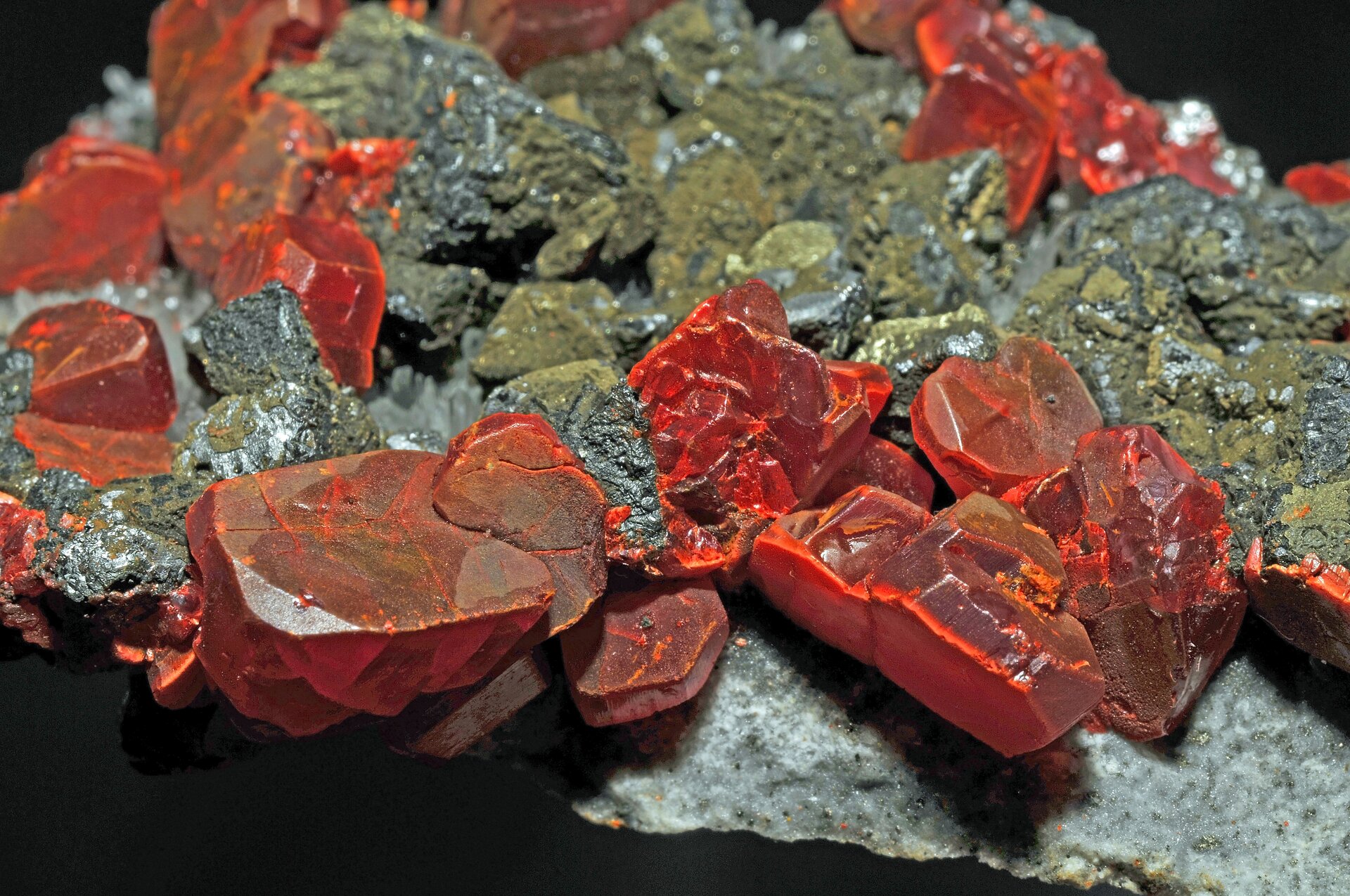 Na zdjęciu jest minerał o pomarańczowoczerwonych kryształach. Kryształy mają kształt wielokątów i liczne zbrużdżenia. Niektóre ścianki są gładkie. Kryształy znajdują się pomiędzy fragmentami innego kamienia.  