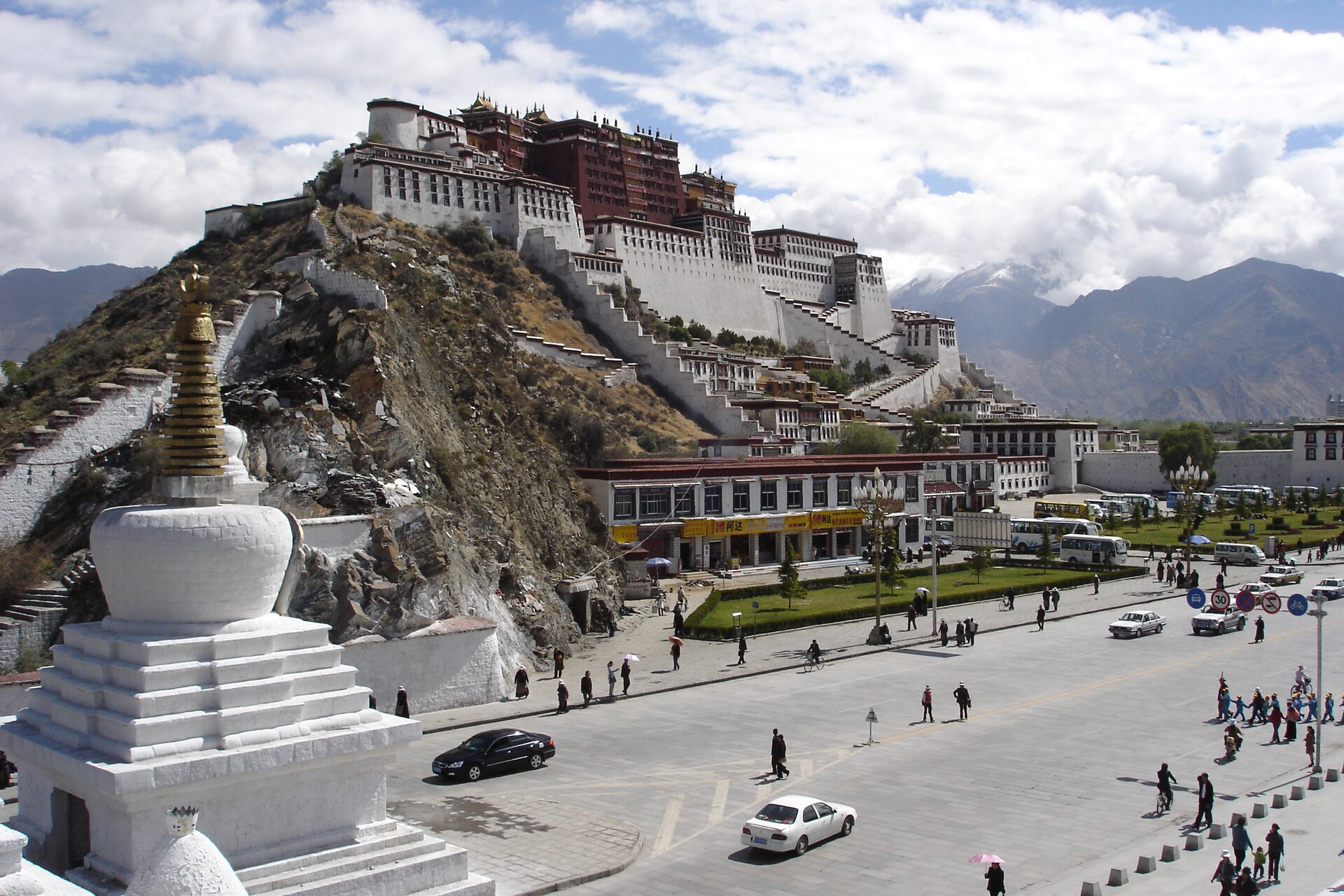 Zdjęcie przedstawia pałac dalajlamy (duchowego przywódcy Tybetańczyków) w Lhasie. Znajduje się on na wzgórzu zaraz przy drodze, jest utrzymany w biało-bordowej kolorystyce. Na placu widoczne są pojedyncze samochody oraz spacerujących ludzi. W oddali widać góry.