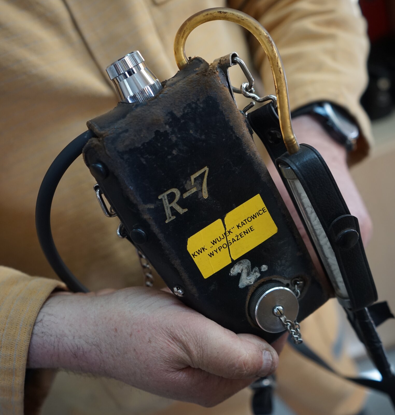 Na zdjęciu widać metanomierz interferencyjny R kreska siedem. Metanomierz jest to przyrząd przeznaczony do ciągłego pomiaru stężenia metanu w atmosferze.Metanomierz widoczny na zdjęciu jest w kształćcie prostopadłościanu. Urządzenie znajduje się w skórzanym pokrowcu. Na dole urządzenia widać srebrną zatyczkę na łańcuszku. Z prawego górnego rogu urządzenia odchodzi rurka. Na przodzie urządzenia znajduje się napis: R kreska siedem. Poniżej przyklejona jest naklejka z napisem: KWK Wujek Katowice wyposażenie.Metanomierz trzyma górnik ubrany w strój roboczy.