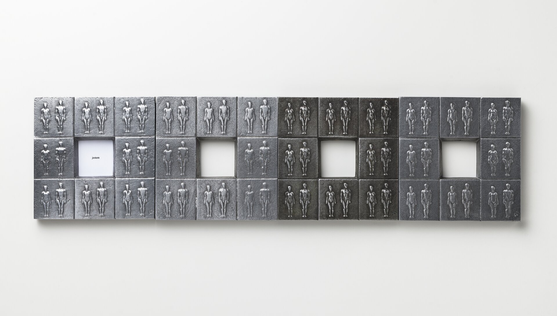 Ilustracja przedstawia pracę Alicji Majewskiej „Tożsamość”. Jest to relief, składający się z 32 metalowych płytek z wizerunkami tych samych, powtarzających się postaci. Praca podzielona jest na cztery kwadraty z czworokątnym otworem w środku. Wszystkie są zespolone w poziomie.