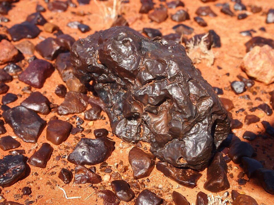 Zdjęcie przedstawia czerwonobrązowe kamienie leżące na powierzchni piasku. Jeden kamień jest większy, pozostałe są mniejsze. Małe kamienie mają gładkie brzegi. Powierzchnia kamieni jest wygładzona, lekko połyskująca w słońcu.  