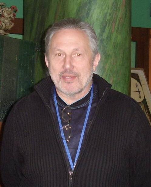 Zdjęcie przedstawia popiersie mężczyzny w średnim wieku. Ma szpakowate, krótkie włosy i kilkudniowy zarost. Jego twarz jest owalna, nos długi, małe oczy osadzone blisko siebie. Mężczyzna ma wysokie czoło. Ubrany jest w zapinany na zamek czarny sweter, na szyi zawieszona niebieska smycz i okulary.