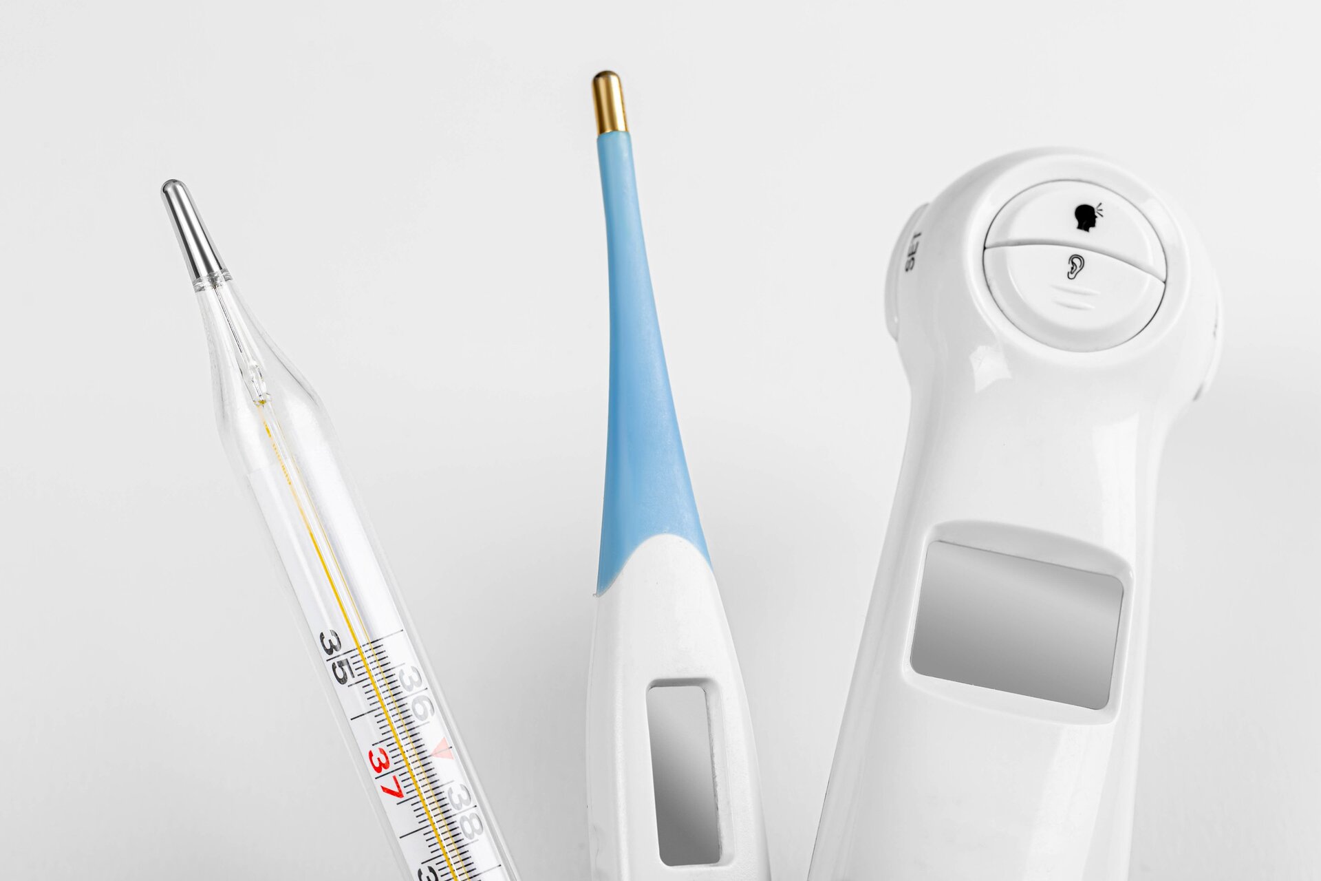 Zdjęcie przedstawia trzy termometry. Widoczny jest termometr cieczowy, który ma wydłużony kształt i rurkę na ciecz w środku. Widać u niego też podziałkę z wartościami. Obok znajduje się podobny do niego kształtem termometr elektroniczny. Posiada on jeden przycisk i ekran wyświetlający wartość temperatury. Obok widoczny jest elektroniczny termometr bezdotykowy. Jest to duży termometr w kształcie litery L. Jedną część zbliża się do czoła, mierzy ona temperaturę. Druga część to uchwyt. Znajdują się na nim przyciski oraz ekran.