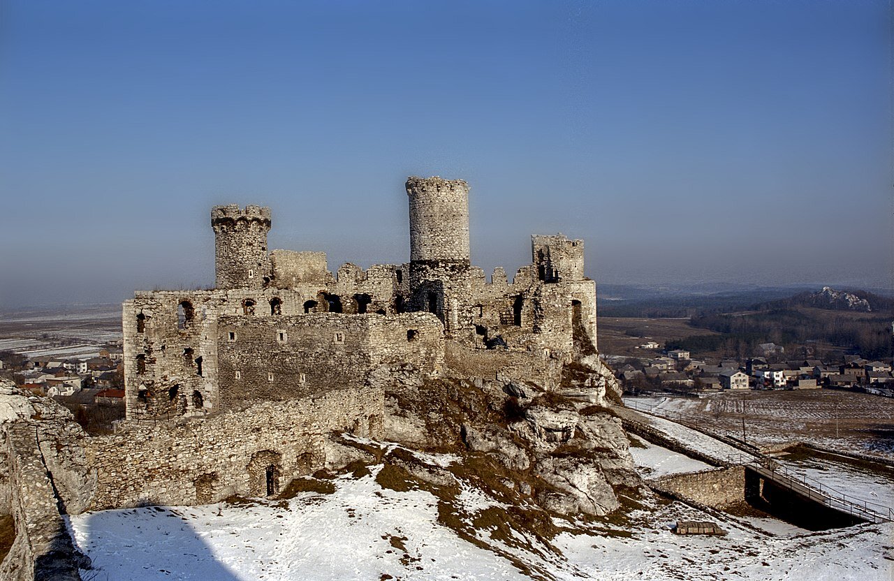 Fotografia przedstawiająca zimowy pejzaż. Pośrodku stoją ruiny gotyckiego zamku wybudowanego z wapiennego białego kamienia. Jest to potężna bryła otoczona skałami. Zamek jest pozbawiony dachu, ma puste otwory okienne. Górują nad nim trzy baszty.