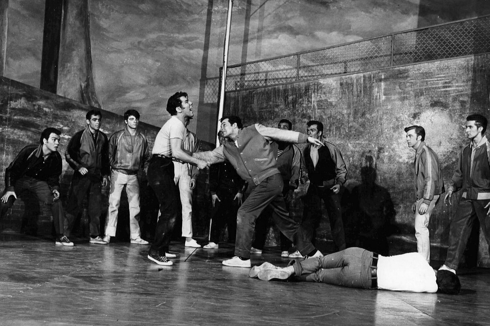 Zdjęcie przedstawia kadr z musicalu „West Side Story”, zdjęcie autorstwa Fred Fehl. Zdjęcie przedstawia grupę młodych mężczyzn. Na pierwszym planie znajdują się dwaj mężczyźni skierowani do siebie twarzą. Mężczyzna po prawej stronie zadaje cios w brzuch mężczyźnie stojącemu naprzeciwko niego.  Po prawej stronie leży bezwładne ciało mężczyzny. Grupa mężczyzn, znajdujących się na drugim planie zdjęcia przygląda się sytuacji. 