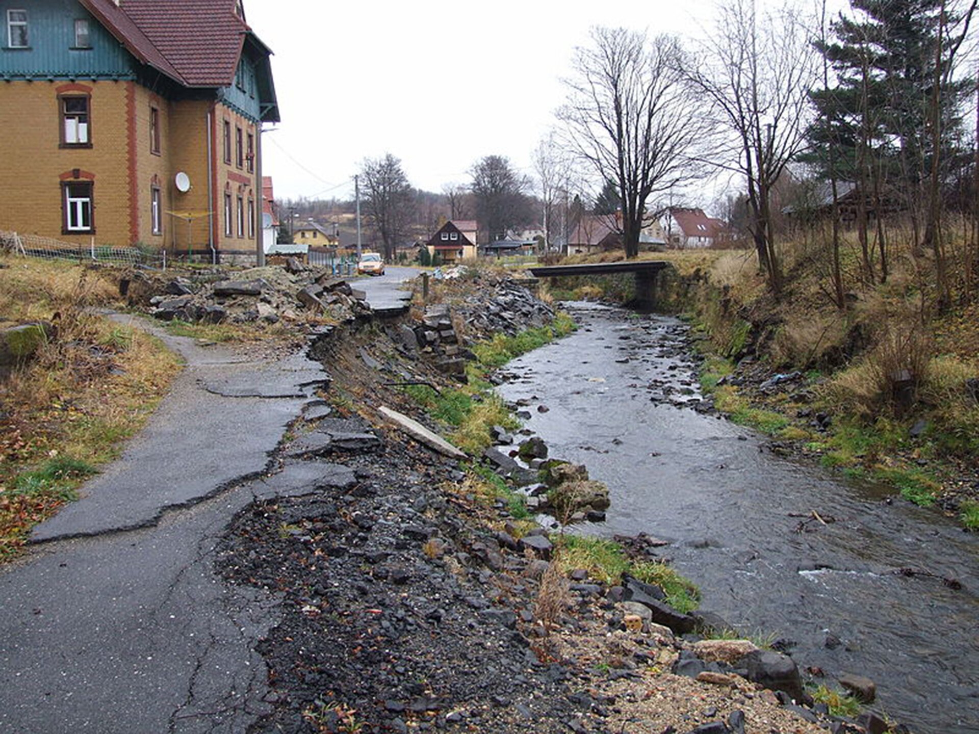 Zniszczenia po przejściu ulewy. Po prawej stronie rwący potok, nad nim mostek i po obu stronach tereny mieszkalne. Z lewej strony zniszczona asfaltowa nawierzchnia drogi znajdująca się bezpośrednio nad płynącą poniżej wodą.