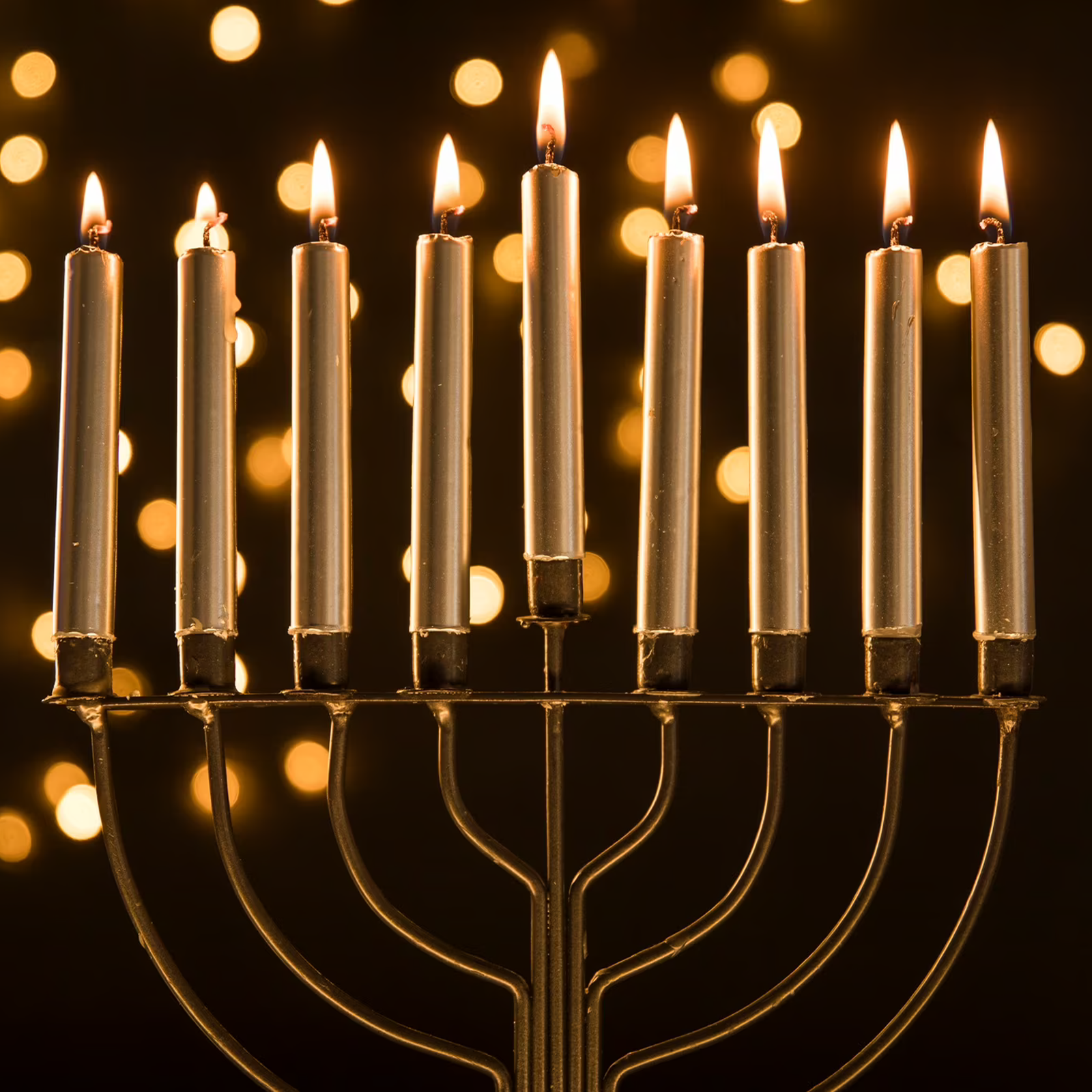 Zdjęcie przedstawia dziewięcioramienny złoty świecznik, tak zwaną menorę, z zapalonymi cienkimi świecami w kolorze kremowym. 