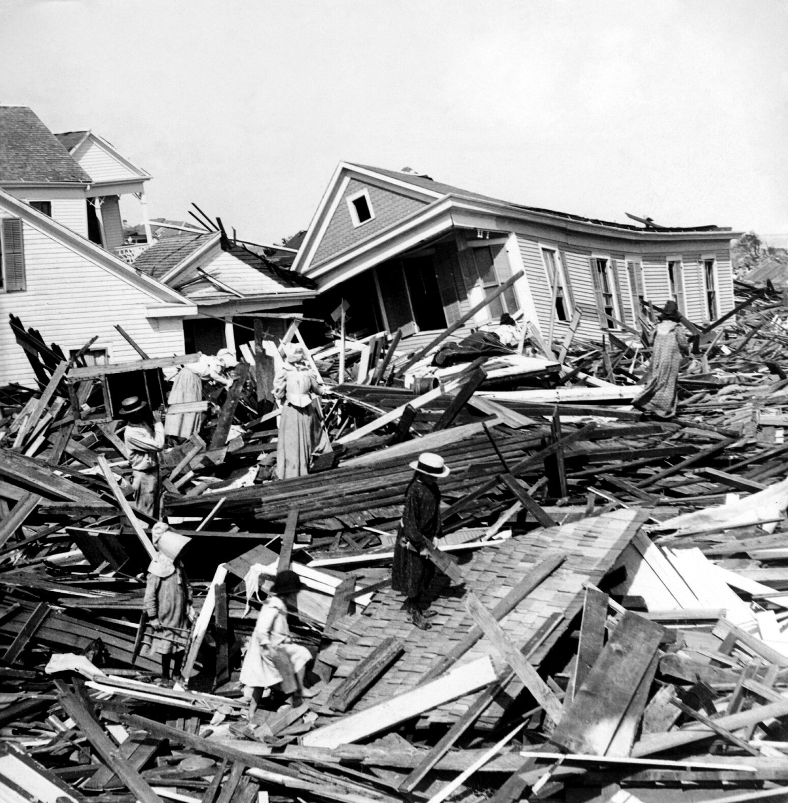 Zdjęcie przedstawia zniszczone drewniane domy. Na pierwszym planie są deski - pozostałości domów. Po deskach chodzą ludzie, szukając czegoś. W tle domy są pochylone, jakby się miały zawalić. 