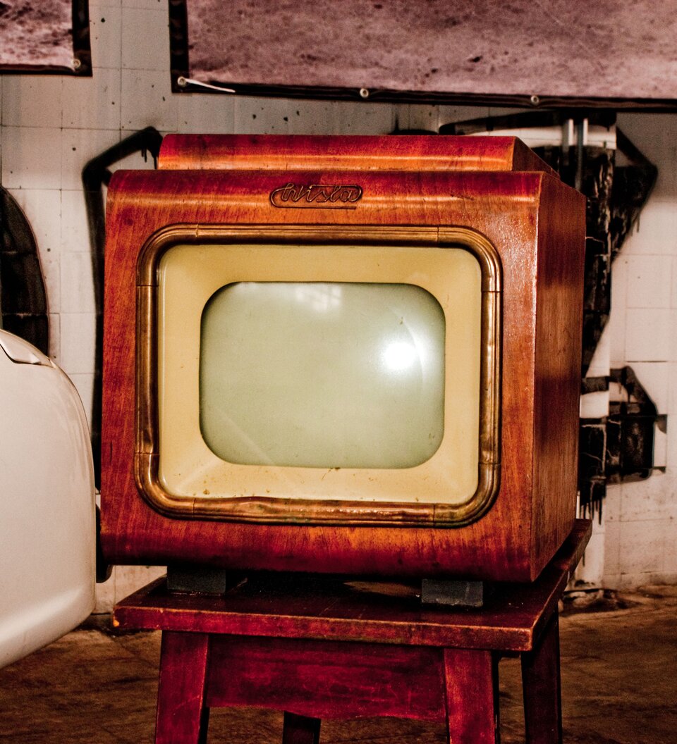 Zdjęcie przedstawia stary telewizor. Ma mały ekran z zaokrąglonymi rogami, który znajduje się w drewnianej, grubej ramie. Na górze ramy jest napis Wisła. Telewizor stoi na drewnianym stoliku.