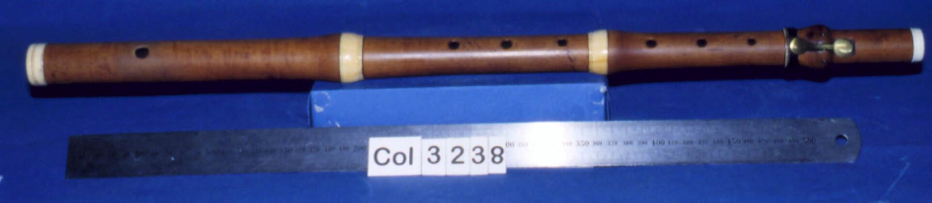 Zdjęcie przedstawia flet barokowy. Ma on prostą budowę - w drewnianej, prostej rurce znajduje się siedem okrągłych otworów. Instrument ma cztery jasne paski, oddzielające kolejne fragmenty. Przy górze instrumentu znajduje się niewielka, metalowa, złota klapka. Przed instrumentem leży metalowa linijka. Flet leży na niebieskim podłożu.