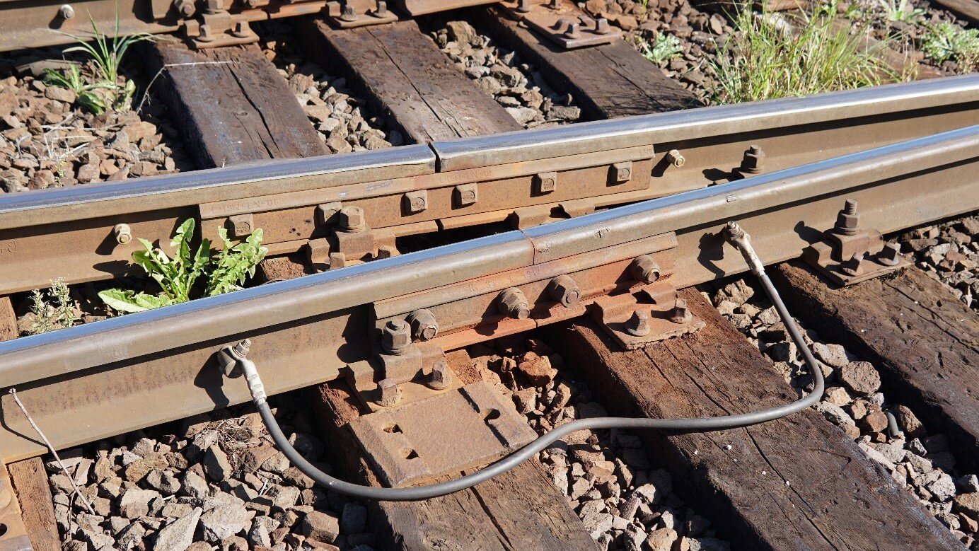 Zdjęcie przedstawia fragment toru. Jest to fragment przedstawiający rozjazd. Widoczna jest szyna zewnętrzna oraz wewnętrzna ułożona pod kątem. Szyny pokazane są w miejscu łączenia dwóch fragmentów szyny. Fragmenty szyny są połączone ze sobą przewodem. 