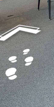 Zdjęcie przedstawia ikonę strzałki i stóp. Na szarej podłodze widoczne są cztery białe odciski butów, powyżej których znajduje się ikona grotu strzałki.
