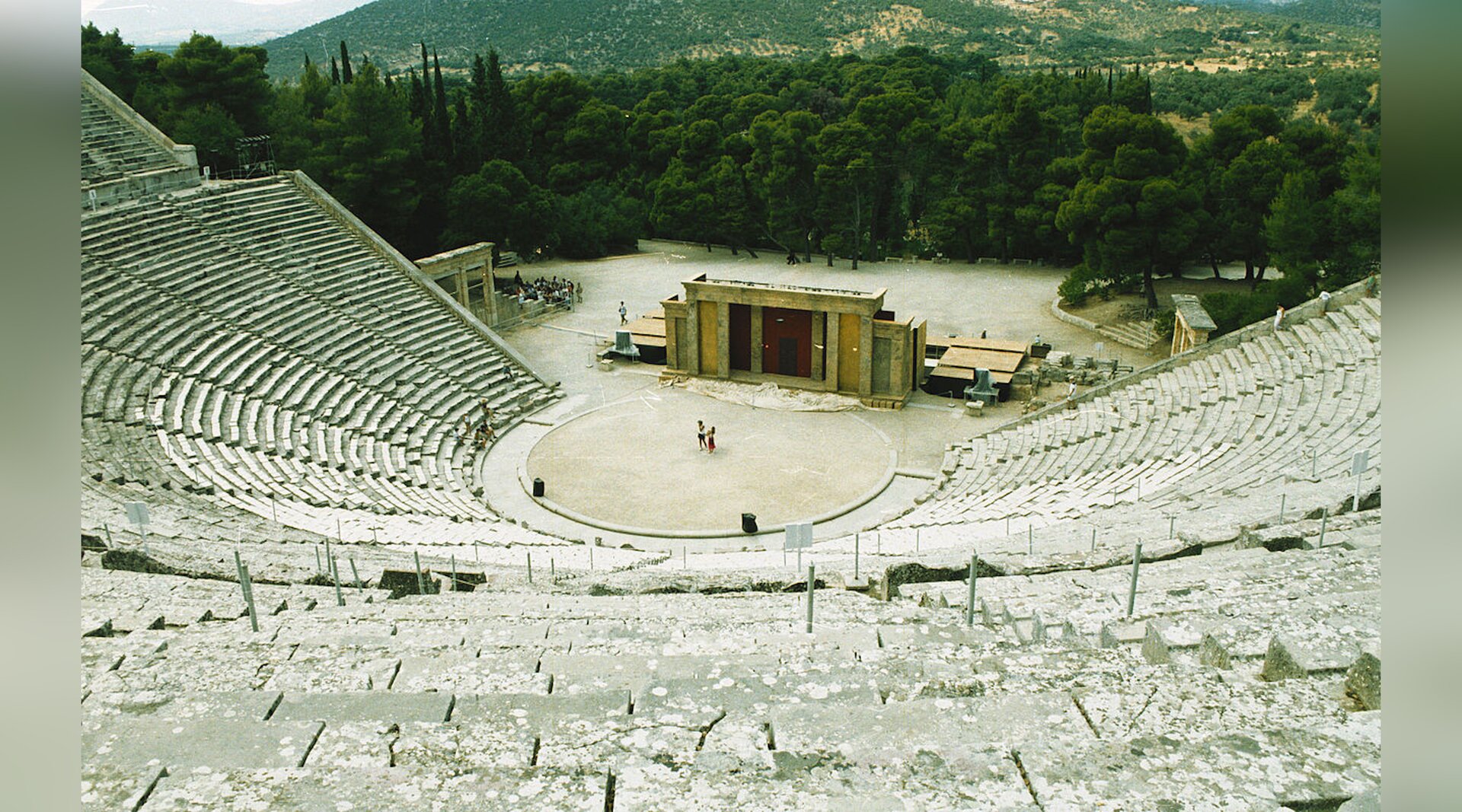 Zdjęcie przedstawia ruiny starożytnego teatru zbudowanego z kamiennych bloków. Widownia znajduje się na zboczach wzniesienia. W tle teatru rośnie las, są zalesione wzgórza
