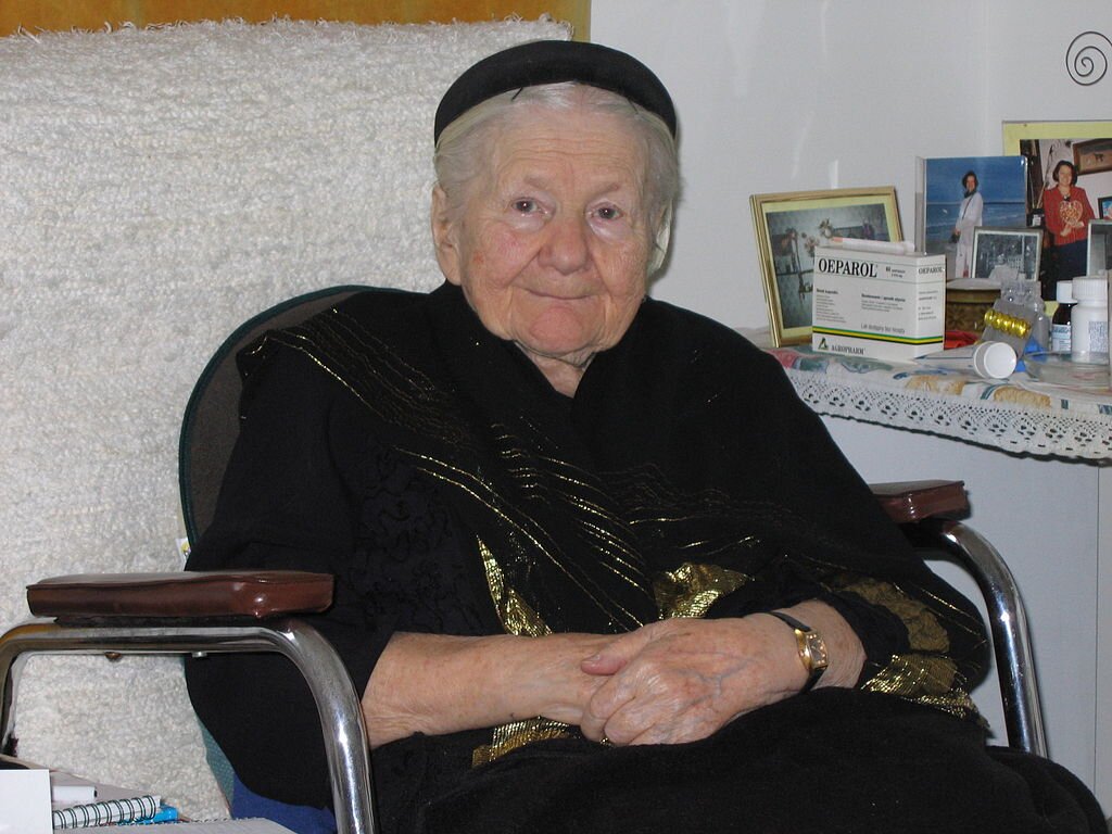 Zdjęcie przedstawia starszą kobietę, która siedzi na fotelu. Ma siwe włosy i czarną opaskę na głowie. Jest ubrana na czarno. W tle po prawej stronie widoczny jest fragment szafki, na której stoją fotografie w ramkach i lekarstwa.