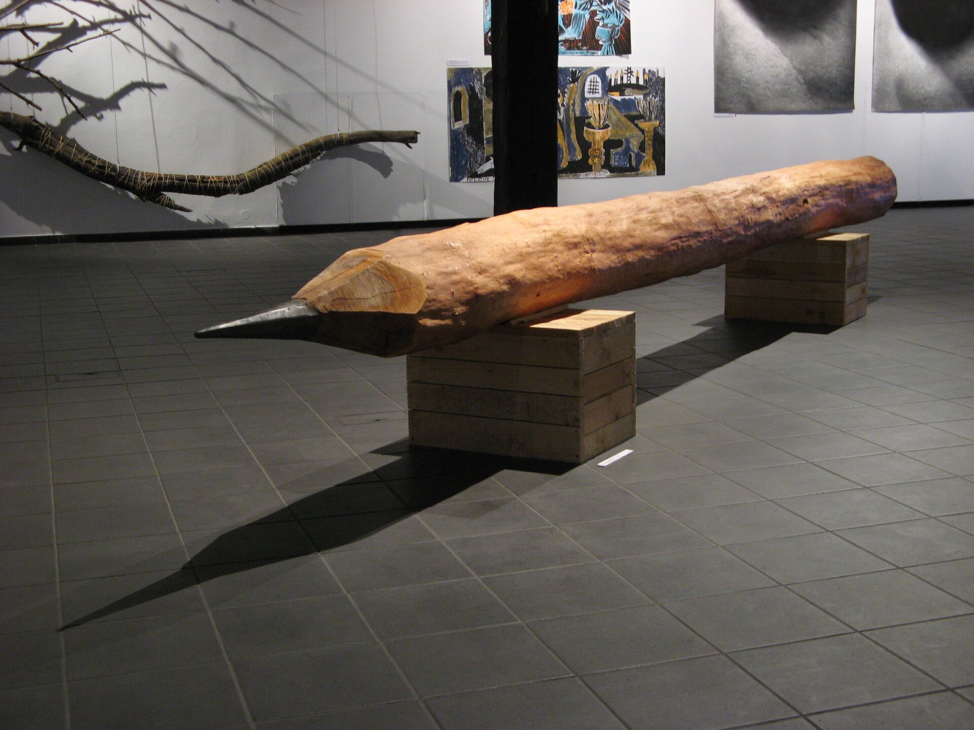 Ilustracja przedstawia salę w galerii sztuki, na środku sali  znajduje się rzeźba „Ołówek” autorstwa Stanisława Kośmińskiego. Rzeźba ma kształt gigantycznego ołówka, który leży na dwóch drewnianych skrzynkach. Ołówek wykonano z kilkumetrowej okorowanej kłody, ma zaostrzony rysik. W tle, na szarej ścianie pomieszczenia umieszczone są prace graficzne oraz malarskie. Po lewej stronie wisi na sznurach, podświetlony duży konar.