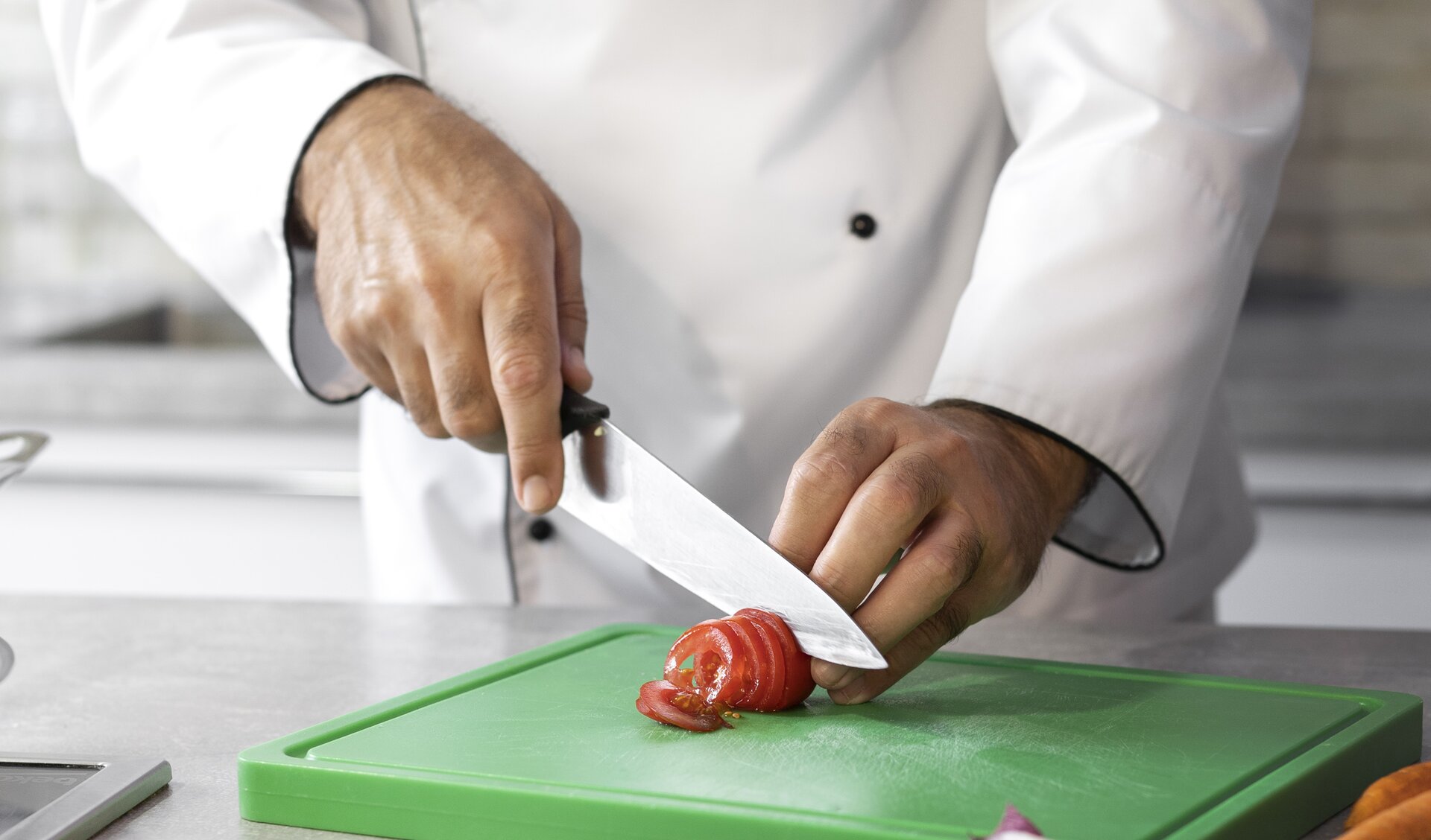 Zdjęcie przedstawia technikę krojenia vichy pomidora. Na pierwszym planie widoczna postać ubrana w strój kucharza, która w prawej ręce trzyma nóż zaciskając palce na rękojeści, którego ostrze  dotyka półplastrów pomidora. Lewa ręka przytrzymuje krojonego pomidora. Pomidor krojony jest na zielonej desce.
