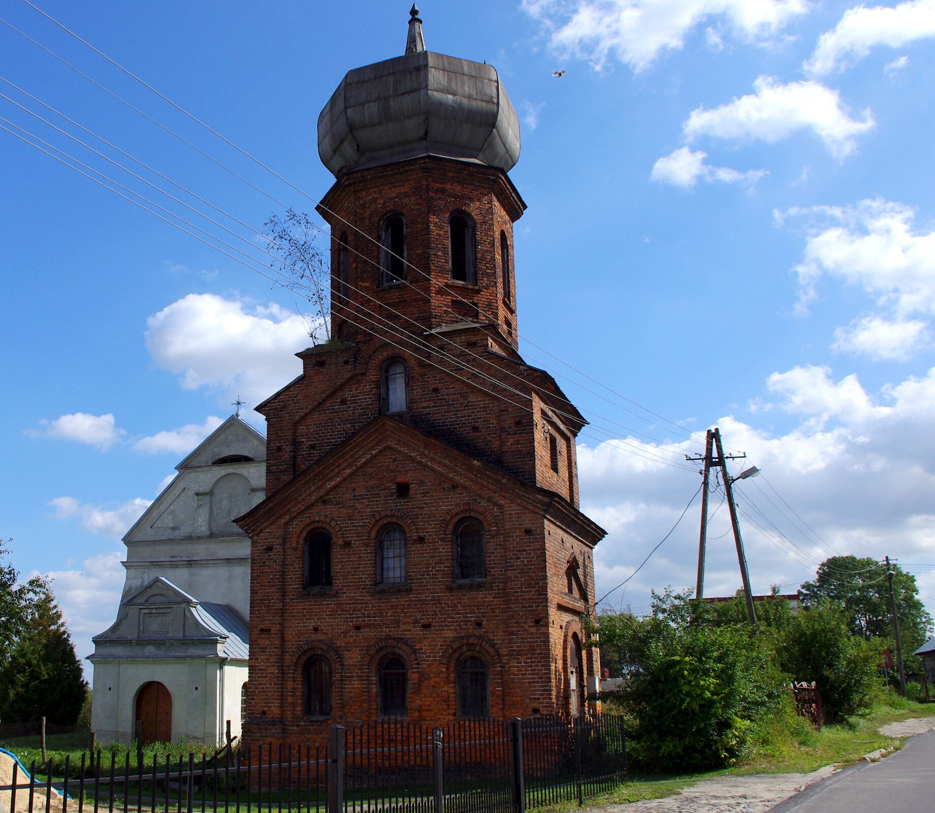  Zdjęcie prezentuje ceglany budynek. Na pierwszym planie wysoka dzwonnica. Zbudowana z czerwonej cegły. W tle niewielka, biała cerkiew.